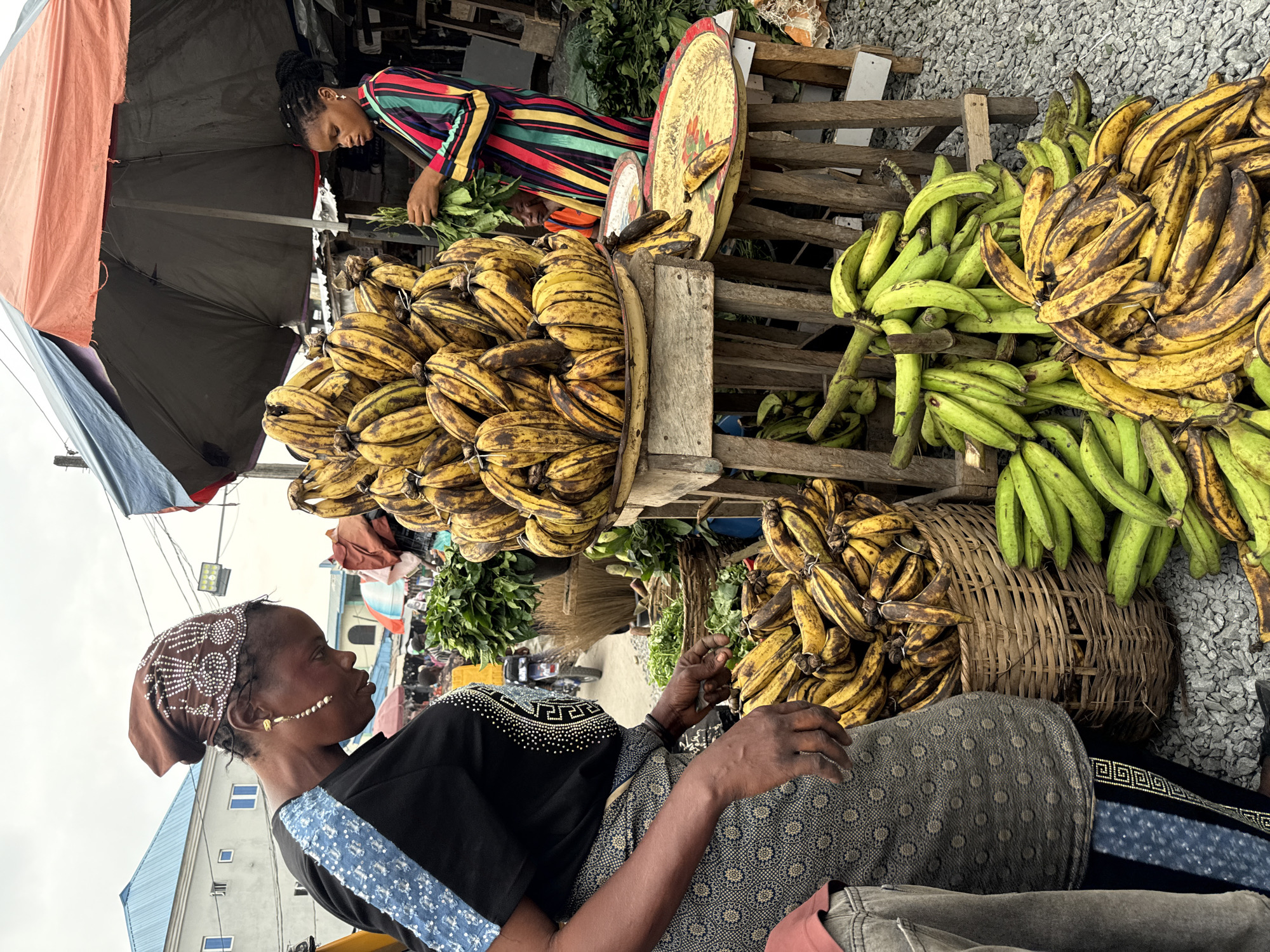 This image captures a vibrant market scene in Ado, Nigeria. A woman in a patterned headscarf and dress sits surrounded by plantains in various stages of ripeness. To her left, a person wearing a colorful striped top is standing behind a wooden structure, likely tending to other produce. Plantains are piled high on wooden crates and in a woven basket. An umbrella provides shade from the sun. In the background, buildings and street signs give context to the urban setting.