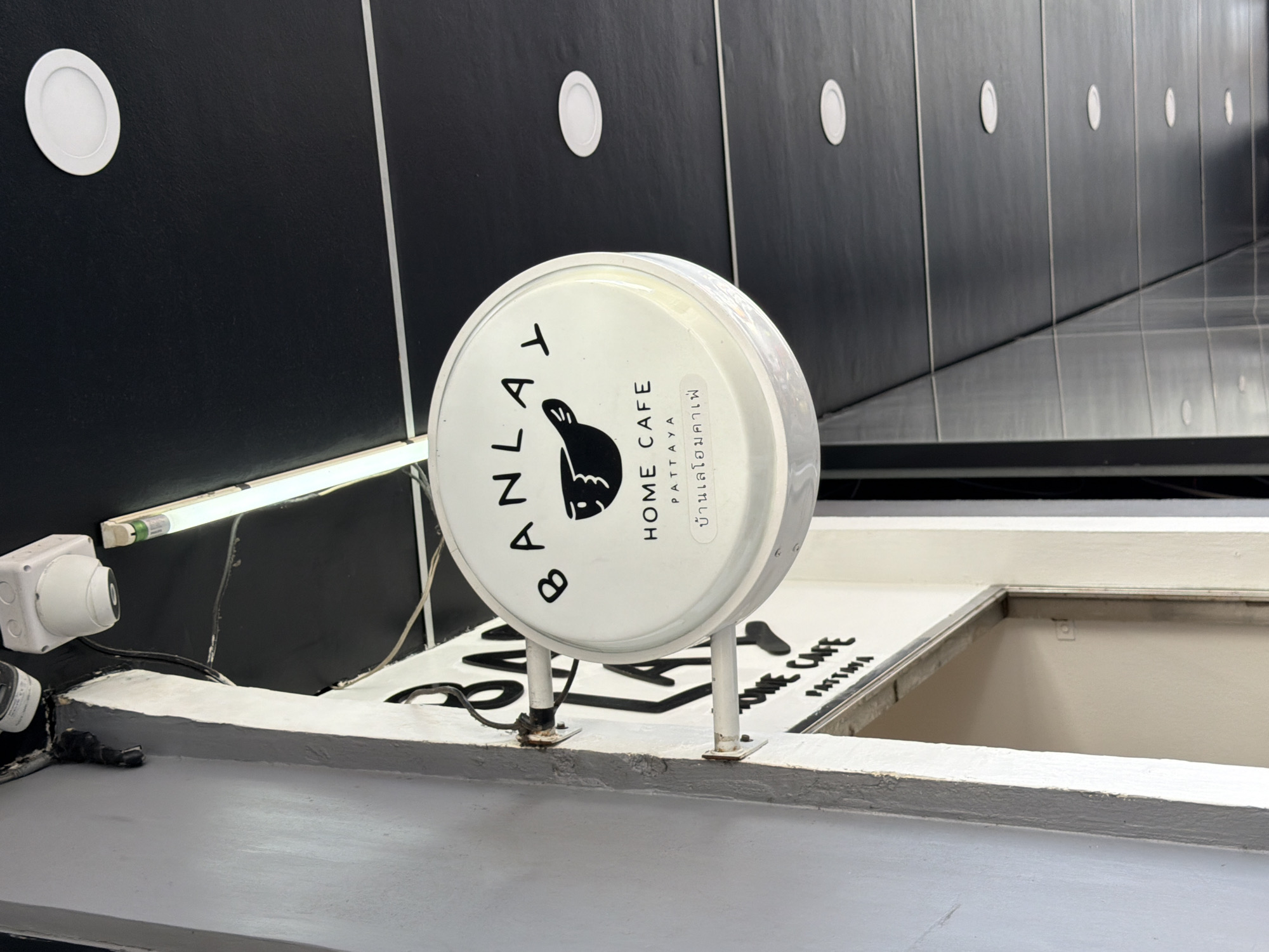 The image shows a circular white sign for "BANLAY HOME CAFE PATTAYA". The sign features a black graphic of a bird and Thai text. The sign is mounted on the exterior wall of a building. Above the sign, a security camera and a fluorescent light fixture are visible. The building appears to be located in an urban environment, possibly a commercial area, given the ceiling with recessed lighting and the presence of a doorway to the right of the sign. The wall to the left of the sign is painted white. The overall scene suggests an establishment in Pattaya City, Thailand.