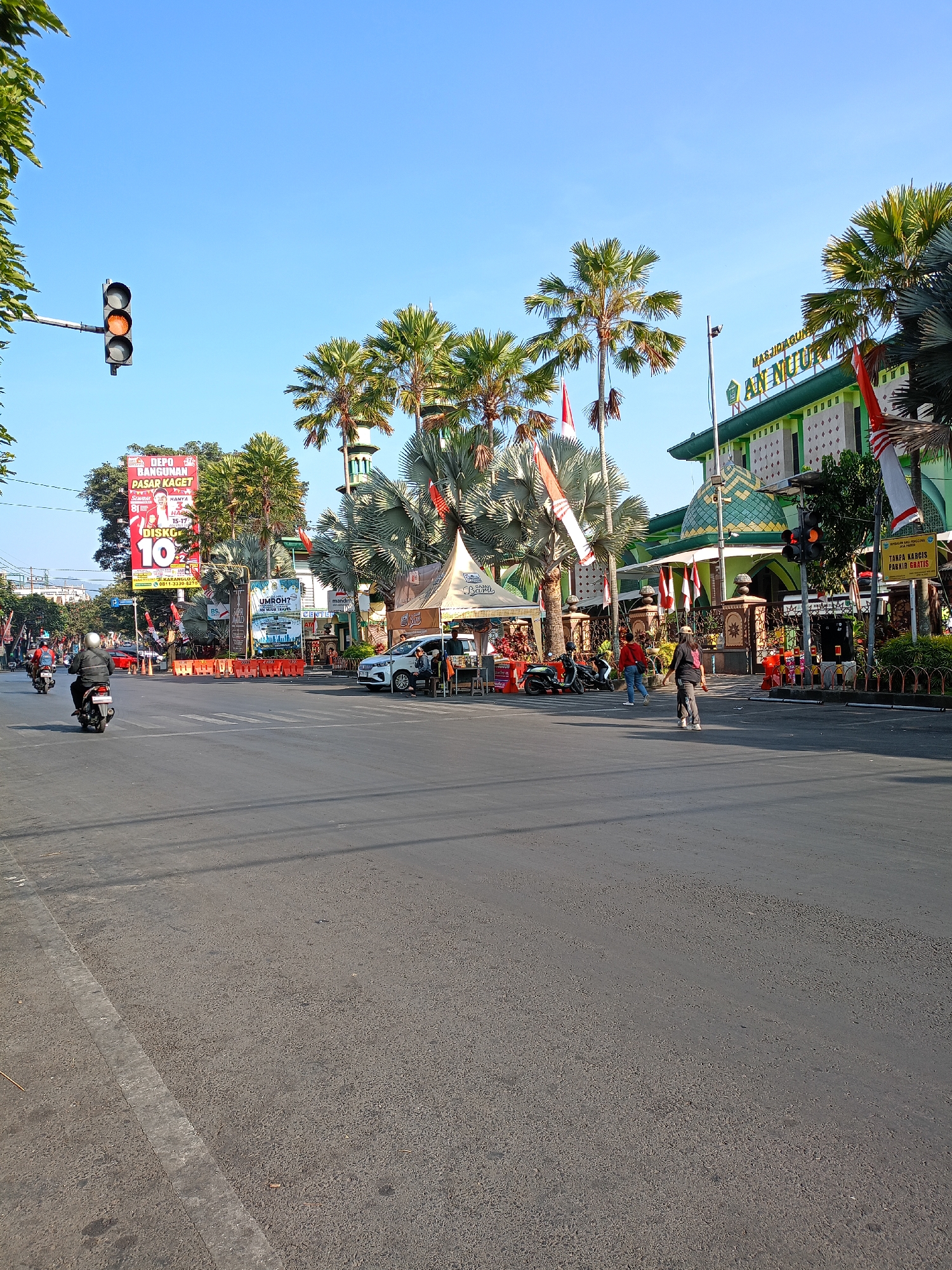 The image captures a street scene in Batu, Indonesia, characterized by a clear blue sky and bright daylight. A traffic light, with its orange signal illuminated, is visible on the left. In the background, the green facade of Masjid Agung An Nur is prominent, adorned with traditional Islamic architectural elements and flanked by lush palm trees. Several red and white Indonesian flags are displayed. Various stalls and tents are set up along the roadside, suggesting a local market or event. People are seen walking and riding motorcycles on the street, indicating moderate activity. The road surface appears worn in some areas, with visible traffic markings. The overall atmosphere is that of a bustling urban environment with a distinct cultural and religious presence.