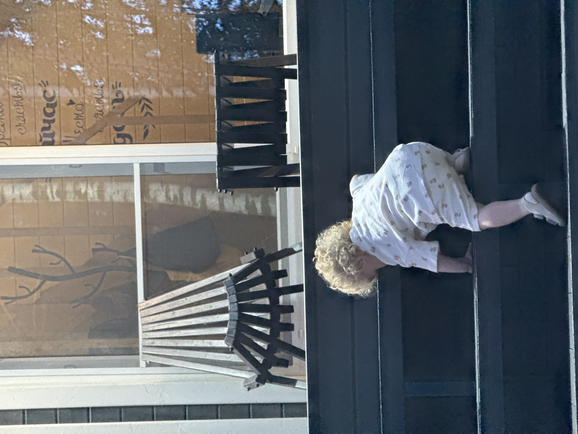 A young child with curly blond hair, wearing a white pajama set with a floral pattern, is climbing up a set of dark gray stairs. The child's back is to the camera, and they are reaching up with their right hand to grasp the next step. The child is wearing white shoes and white socks. The stairs are outdoors, and in the background, there is a wooden building with a window. A coat rack with branches is visible through the window, along with some text written on the building's exterior. There are also two dark wooden benches or seating structures in the background. The scene appears to be in Prokudino, Russia.