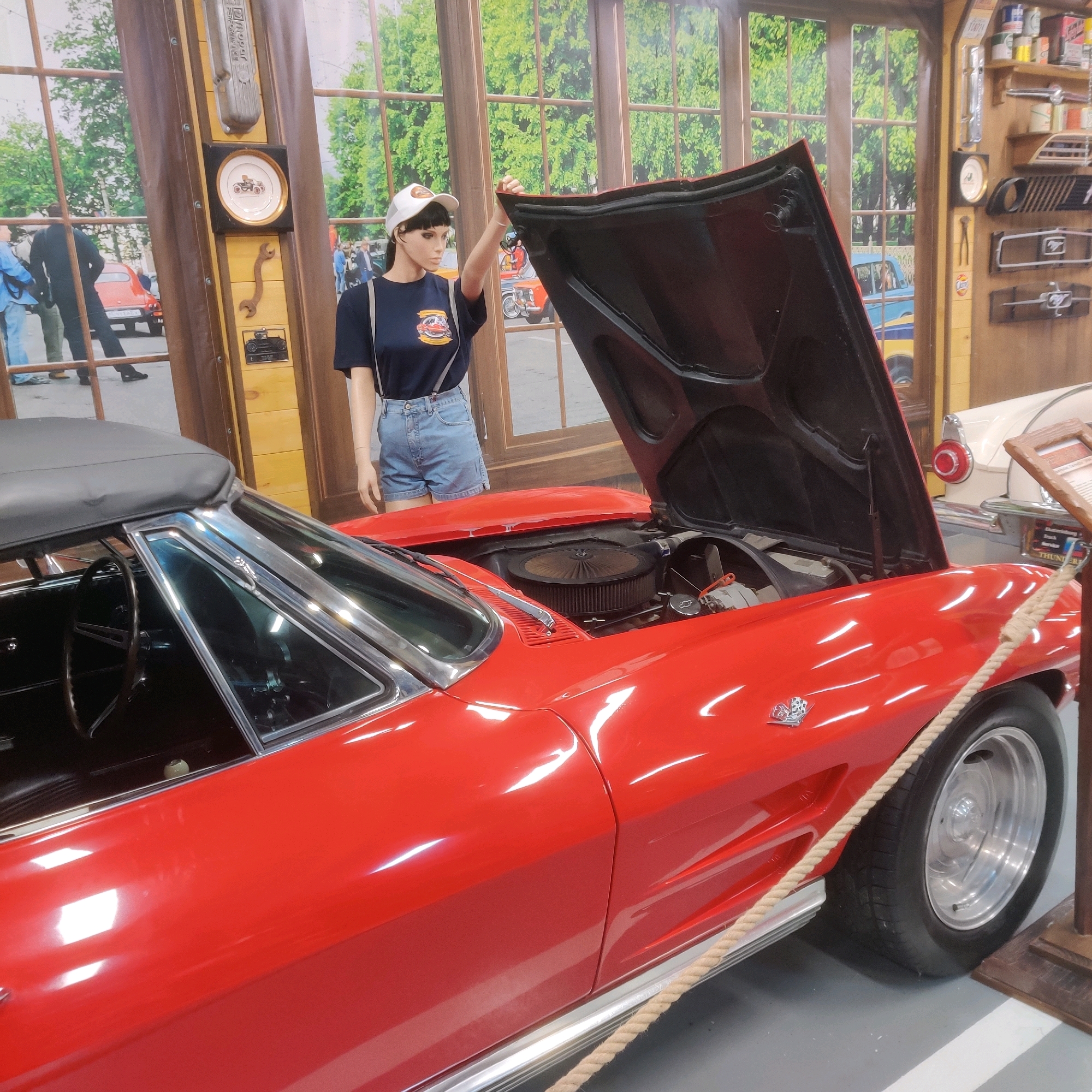 This image depicts a red convertible car, possibly a Chevrolet Corvette, on display in what appears to be a museum or exhibition. The car's hood is open, revealing the engine compartment. A mannequin dressed in a blue t-shirt, denim shorts, and a white baseball cap stands beside the car, with its hands near the open hood. The background features a mural of a street scene with trees and buildings, and shelves adorned with car parts and memorabilia. The lighting is bright, illuminating the polished surface of the car. The image was taken in Yekaterinburg, Russia.