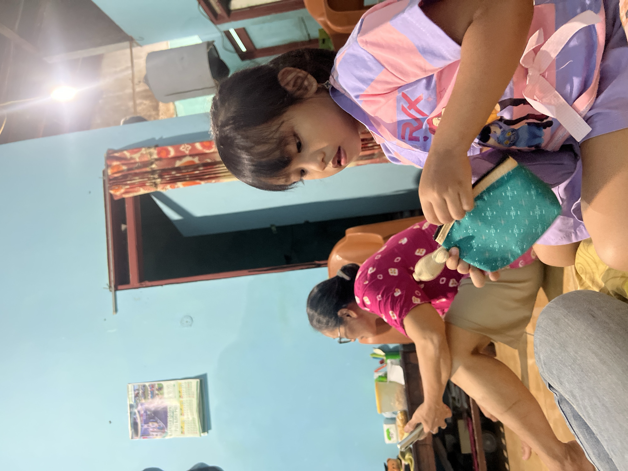 A young child wearing a pink and purple striped shirt and shorts is focused on a small teal pouch with a patterned design. The child is looking down, with their mouth slightly open, seemingly engaged in an activity with the pouch. Behind the child, another person, an adult woman wearing glasses and a pink shirt with a white circular pattern, is leaning forward and also appears to be interacting with something. The adult's hair is pulled back into a bun.
The scene takes place indoors, with light blue walls visible. A patterned curtain hangs in a doorway to the left. A calendar with images is attached to the wall on the left side of the frame, and there is a bright light source emanating from the ceiling on the upper left. The overall impression is of a casual, domestic setting. The image appears to be taken in the daytime, given the lighting. There is no information about the weather or specific emotions conveyed beyond the child's focused expression. No discernible text is visible within the image, other than potentially on the calendar, which is not clear enough to read. The overall orientation of the image is rotated. The location is Kecamatan Pati, Indonesia.