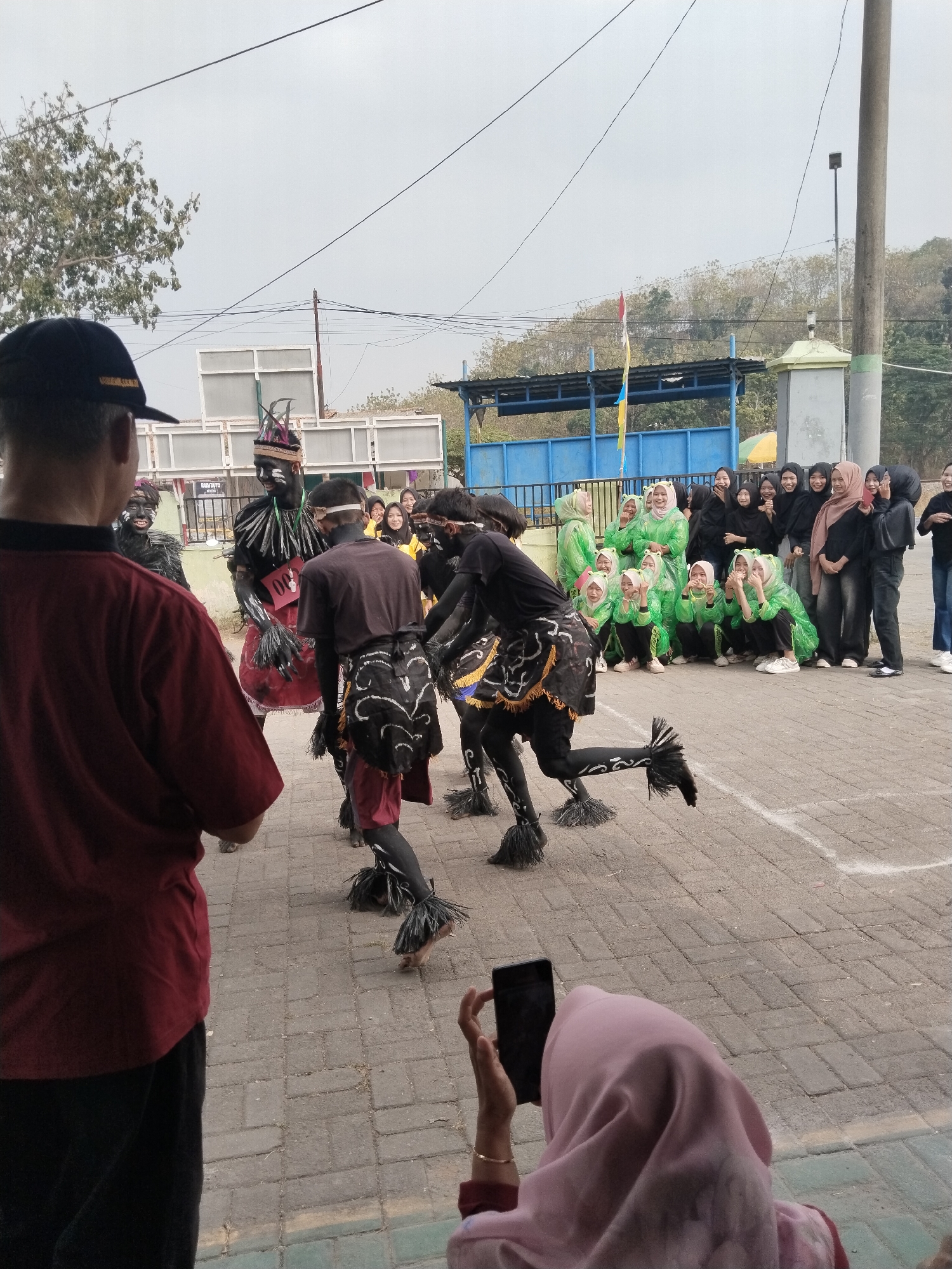 In Pakulaut, Indonesia, a street performance unfolds on a paved area. A group of individuals in black and red costumes, adorned with tribal-like face paint and headdresses, are engaged in a dance. One performer, centered in the frame, wears a dark headdress with feathers and a red skirt, and a badge with "06" is visible. Two other performers in similar attire are to his left, one with a dark headdress and face paint, and the other with a dark shirt and red shorts. To the right, a person in a maroon shirt faces away from the camera. In the background, a group of onlookers, predominantly young women wearing green and black hijabs, watch the performance. A person in a pink hijab is in the foreground, capturing the event with their phone. The environment includes a blue structure with a roof, trees, and a grey pole with a light fixture. The sky is overcast.
