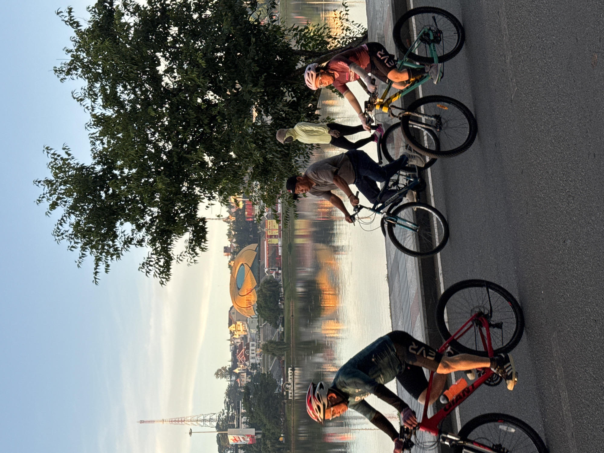 The image depicts a scenic view of Xuan Huong Lake in Da Lat, Vietnam, during a golden hour sunrise. In the foreground, four individuals are cycling along a paved path beside the lake. The first cyclist from the left is wearing a teal long-sleeved cycling jersey and black cycling shorts, with a red and white helmet. The second cyclist is a man in a grey t-shirt, dark pants, and a dark baseball cap, riding a blue bicycle. Behind him, a person in a light yellow hoodie and black pants is walking. The fourth cyclist, on the right, is a woman wearing a burgundy long-sleeved top and black cycling shorts, with a pink helmet.
The lake occupies the mid-ground, reflecting the warm hues of the sunrise and the surrounding cityscape. Across the lake, the Da Lat city skyline is visible, featuring a distinctive golden-domed building, residential houses with red roofs, and a tall radio tower on the left. The sky is a clear blue with a few wispy clouds, and the overall atmosphere suggests a peaceful morning activity. The lush green foliage of a large tree is prominent on the right side of the image, framing the scene.