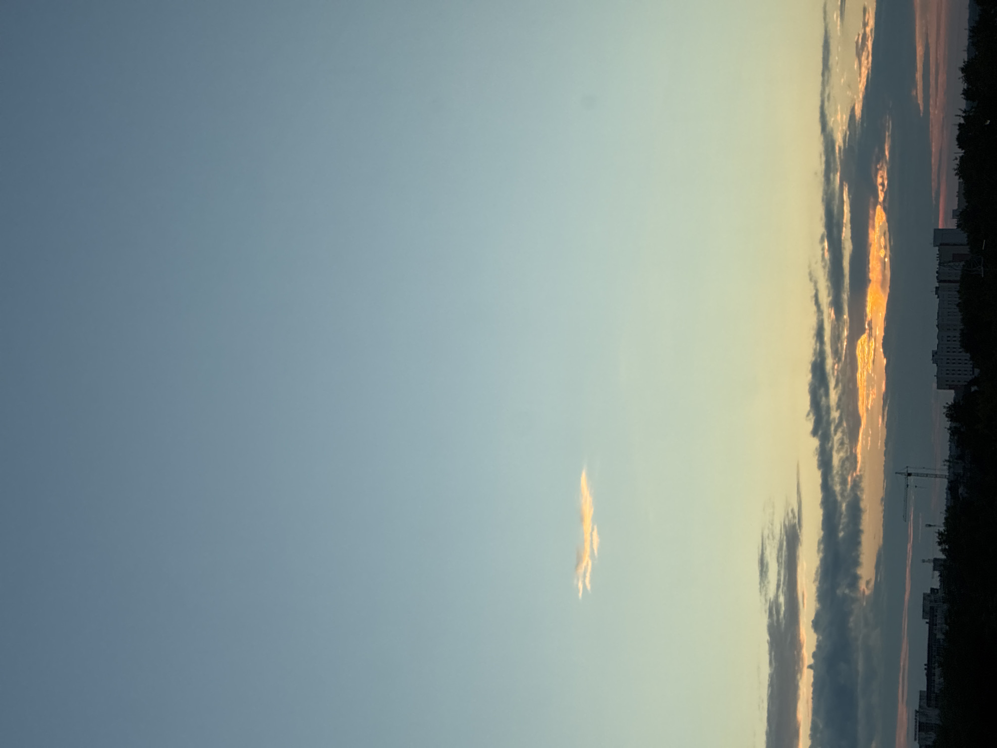 The image captures a serene sky over a city at dusk. The sky transitions from a pale blue at the top to a soft yellow and orange hue closer to the horizon. A few scattered clouds are visible; a small, wispy cloud tinged with yellow floats in the upper middle section of the frame, while a band of darker, more textured clouds runs across the lower portion of the sky, catching the warm light of the setting sun.
Below the horizon, the silhouette of a cityscape is visible. Buildings of varying heights and architectural styles are discernible, some appearing to be apartment blocks or office buildings. Amidst the urban landscape, a construction crane stands tall, suggesting ongoing development. Dark green foliage, likely trees, forms a dense line along the base of the cityscape, softening the urban edge. The overall impression is one of a peaceful transition from day to night in an urban environment.