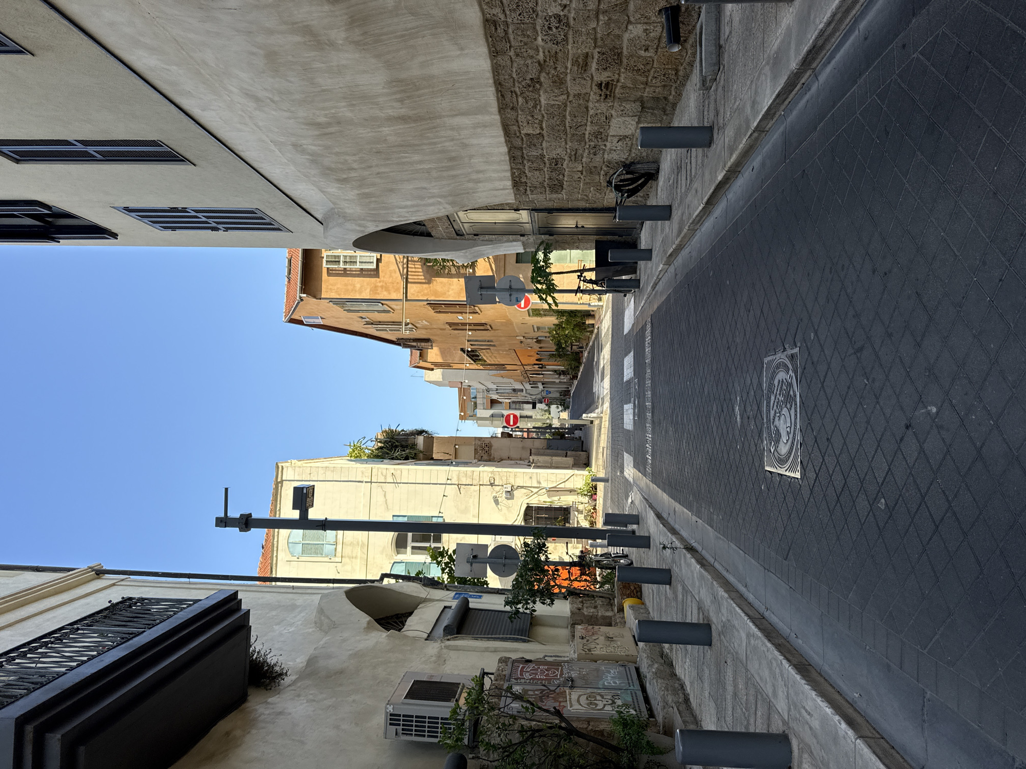 The image shows a narrow street in Tel Aviv, Israel, on a clear, sunny day. The street is paved with dark gray tiles in a diamond pattern, and lined with buildings on both sides. The buildings are of varying heights and architectural styles, with some featuring traditional stone facades and others with modern stucco finishes. Many of the buildings have balconies with decorative iron railings, and windows with shutters.

The street appears to be a pedestrian walkway or a quiet street with limited vehicle traffic, as evidenced by the lack of cars and the presence of bollards along the sidewalk. There are some potted plants and trees along the street, adding a touch of greenery.

Notable details include several gray cylindrical bollards spaced along the street, a metal grate embedded in the pavement, and a red and white circular traffic sign. One building has an air conditioning unit mounted on its exterior wall, and another has graffiti on its lower section. In the distance, the street curves and narrows, suggesting it leads to a more densely built area. The overall impression is one of an old, charming urban environment bathed in bright sunlight. There is no visible text of significance in the image.