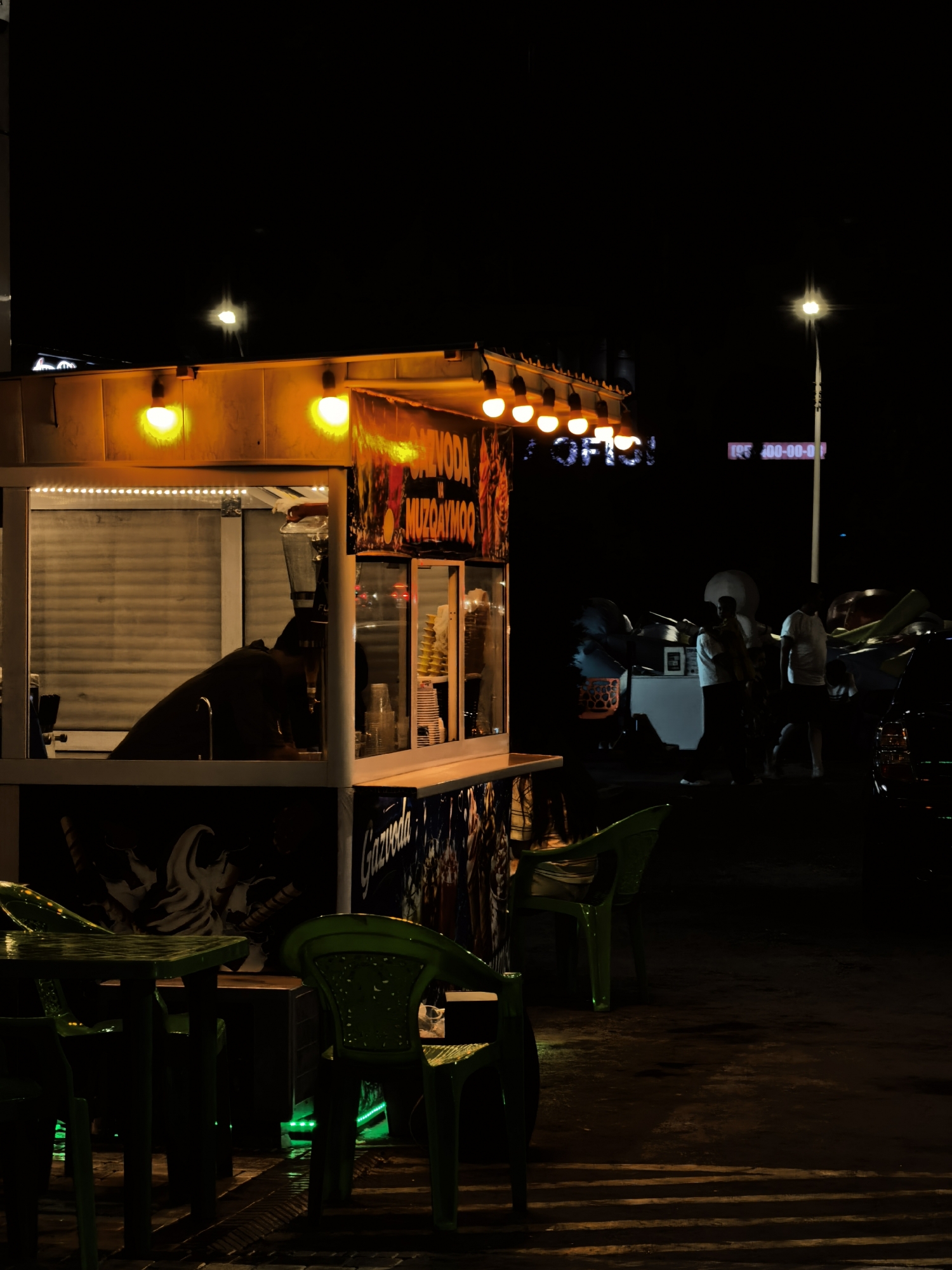 An outdoor food stall is depicted at night in Fergana City, Uzbekistan. The stall, illuminated by orange overhead lights, features a serving window where a person in dark clothing is visible behind a blender. Advertisements for "GAZOVODA MUZGOYMOQ" are displayed on the stall's exterior. In front of the stall, several green plastic chairs and tables are arranged on a wet, paved surface, reflecting green and orange lights. In the background, several individuals are gathered near other stalls and vehicles, illuminated by a streetlamp. The overall scene is dark, with artificial lighting dominating the environment.