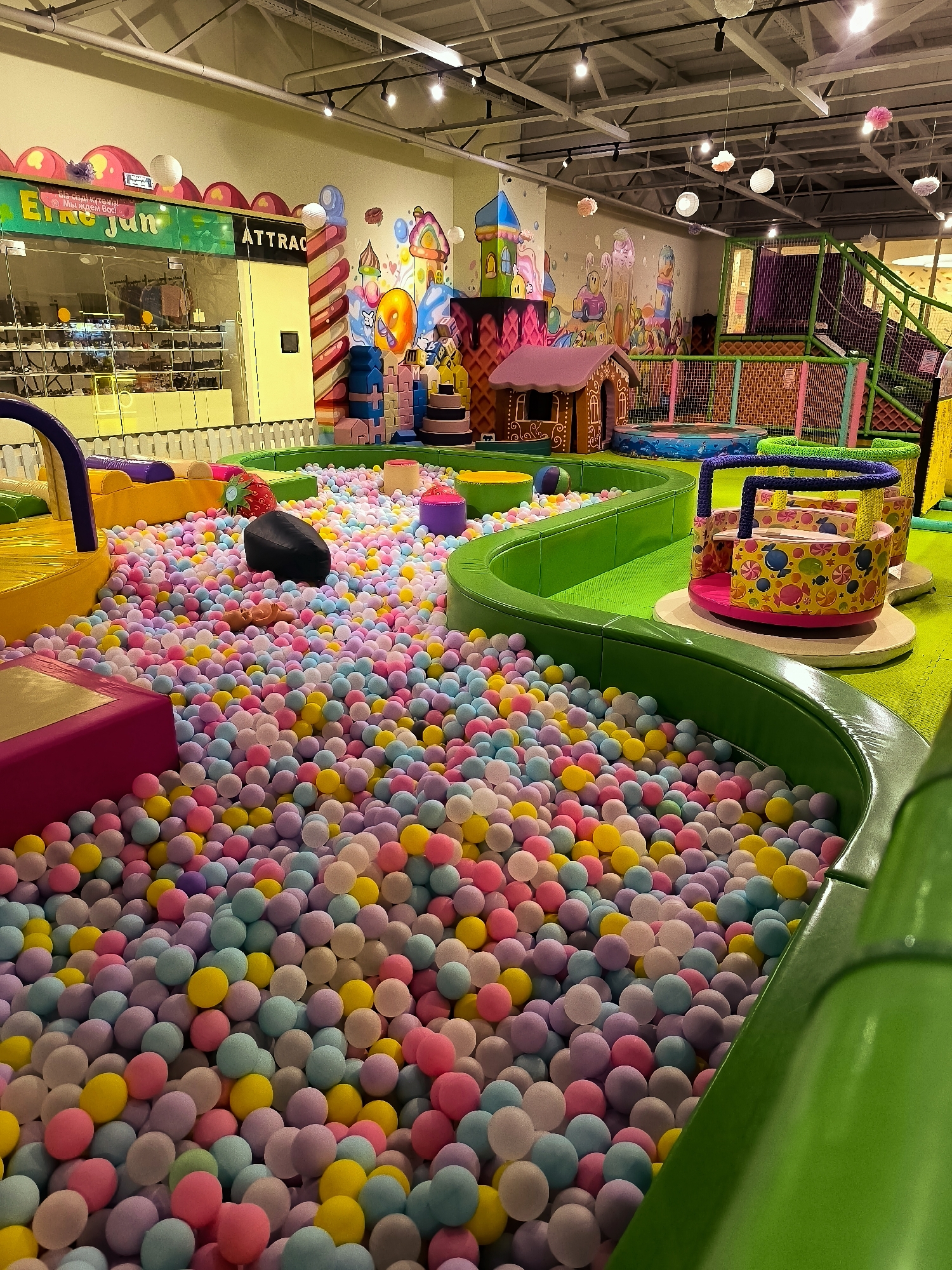 The image shows a brightly colored indoor playground for children, likely located in the city administration area of Qarağandı, Kazakhstan. The main feature is a large ball pit filled with a multitude of pink, yellow, blue, and purple balls. Surrounding the ball pit are various play structures, including green padded slides and ramps, a small carousel with a candy-themed design, and a trampoline. In the background, there's a wall mural depicting a whimsical candy land with castles and sweets, and a section of what appears to be a store or reception area. The overall environment is clean and well-lit, suggesting a safe and fun place for children to play. There are no people visible in the image.