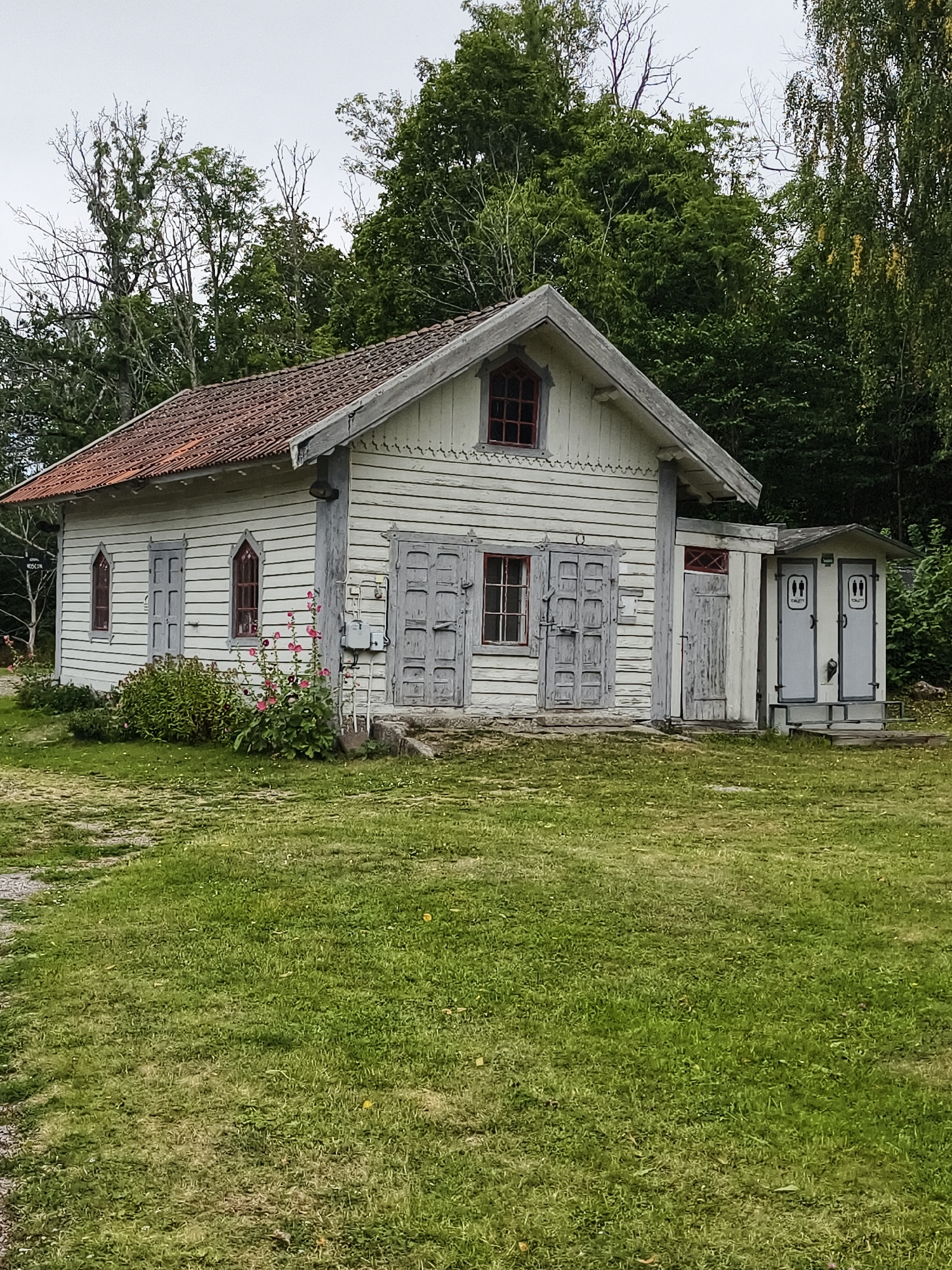The image shows a small, white wooden building with a red tile roof, located in Brokind, Sweden. The building appears to be old, with weathered clapboard siding and decorative trim under the eaves. There are several windows with red frames and dark panes. Two large double doors are visible on the front of the building, as well as a smaller door to the right. Adjacent to the main building is a small, attached structure that appears to be a public restroom, with two separate stalls, each with a door displaying a "toilet" symbol. The building is surrounded by a grassy lawn and a backdrop of mature trees. The sky is overcast. There are no people visible in the image.