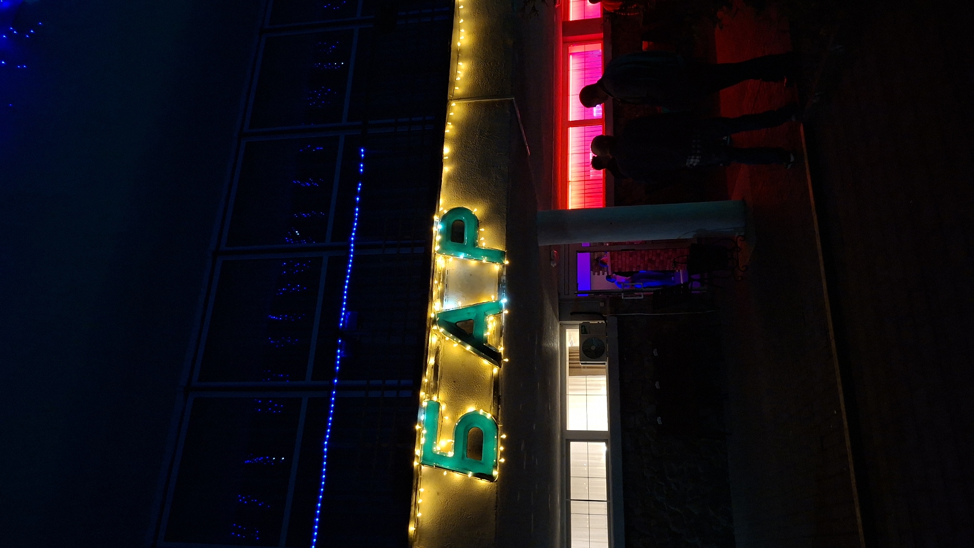 At night in Нарацкі сельскі Савет, Belarus, the vertically oriented image shows the illuminated sign for a "BAR" in bright green letters outlined with warm yellow lights. The sign is attached to a light-colored vertical wall. Blue lights are visible on a darker building to the left. Silhouettes of two people are seen near red-lit windows to the right. The foreground is dark.
