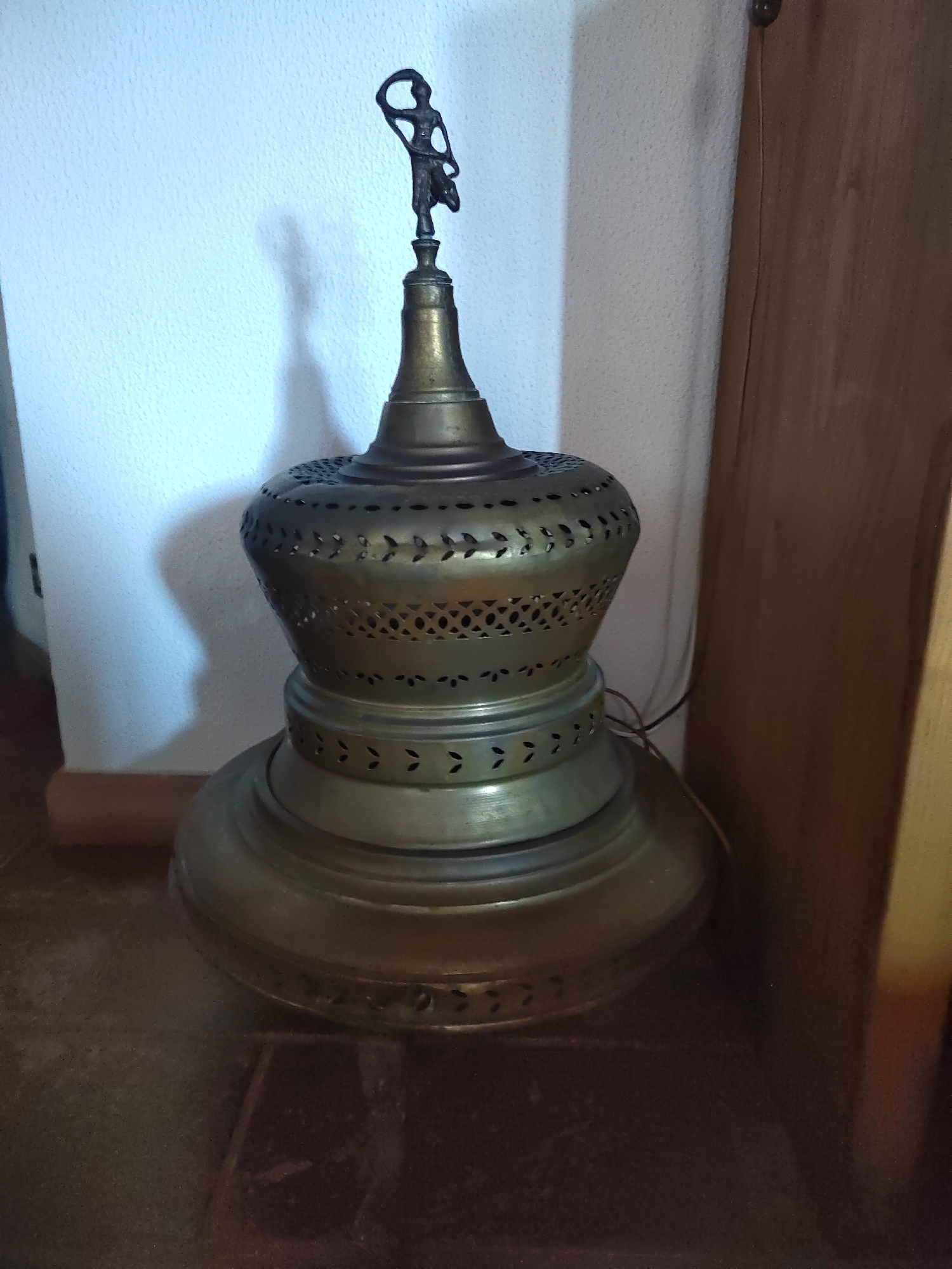 An antique brass lamp with intricate cutouts and a decorative finial depicting a figure with a hoop stands on a dark floor. The lamp is positioned against a textured white wall, with a hint of a wooden surface visible to the right. The scene appears to be indoors, likely in a domestic setting. There are no people or animals present in the image, and no specific identifiable landmarks within Porto Recanati, Italy are visible.