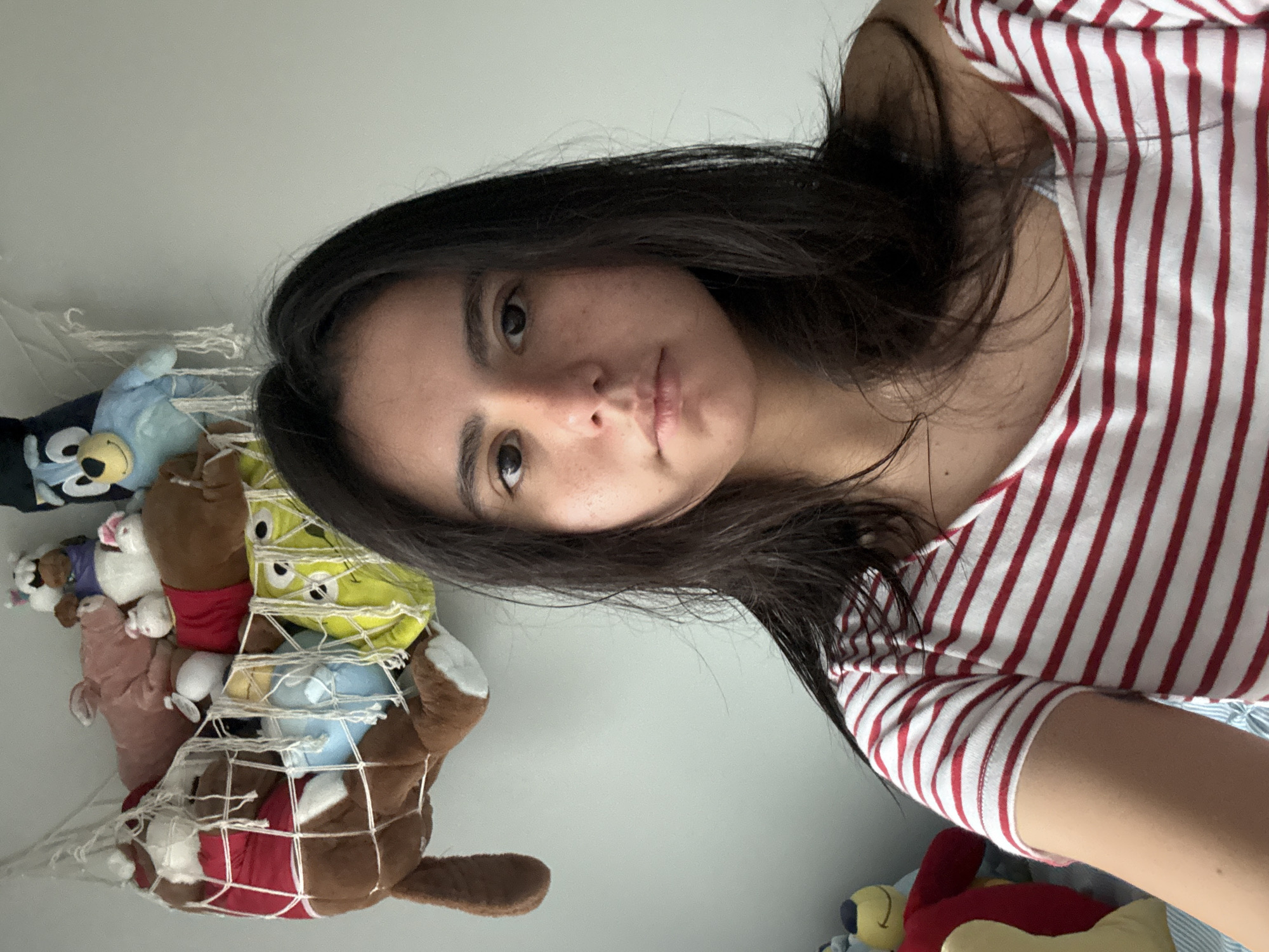 A young adult female is the main subject of the image, wearing a red and white striped shirt. Her dark hair is shoulder length. She is taking a selfie indoors, with a white wall as the background. A net filled with various stuffed animals is visible to the left. The location is not discernible from the image, so an association with the city of Austin is speculative.