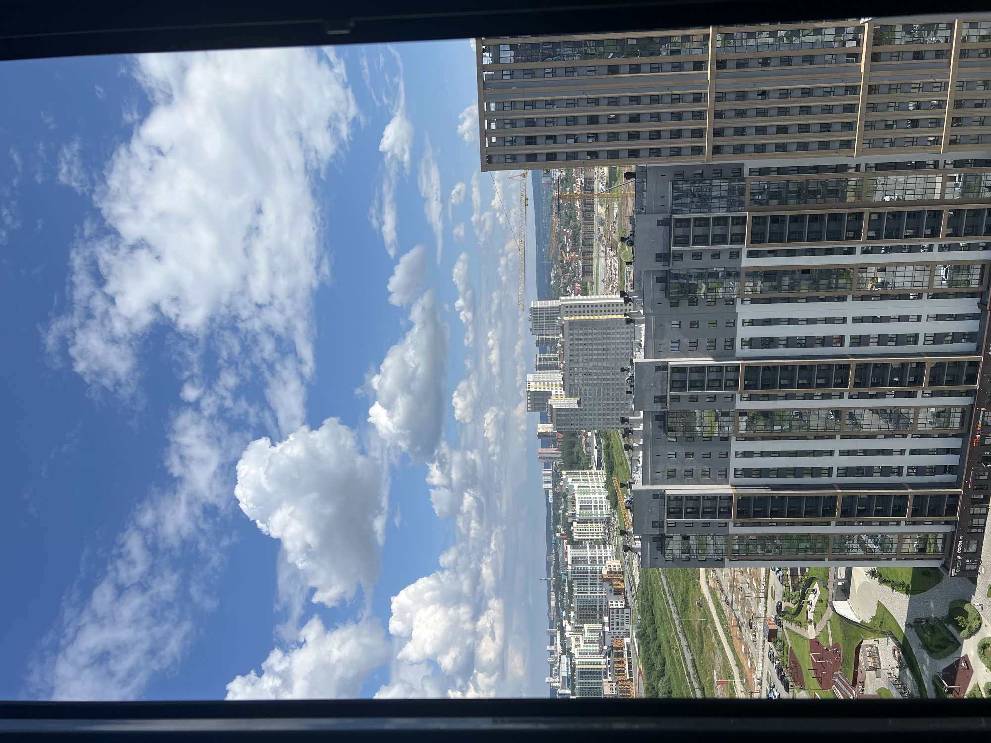 The image captures a panoramic view of a contemporary urban landscape in Yekaterinburg, Russia, under a bright, cloud-dappled sky. In the foreground, two modern high-rise apartment buildings dominate the scene. The building on the left features a striking facade with alternating bands of grey and white, interspersed with vertical panels of reflective glass. Its lower section appears to be a parking lot with parked vehicles and a children's playground with red and yellow play structures, surrounded by green lawns and paved walkways.
The building on the right is taller and exhibits a more uniform design, with a beige exterior and regularly spaced windows. Between these two prominent structures, a network of roads and green spaces is visible, with some areas appearing to be under construction, indicated by the presence of a yellow construction crane in the distance.
Further in the background, a sprawling cityscape unfolds, characterized by a multitude of residential and commercial buildings of varying heights and architectural styles. The cityscape extends to the horizon, where a faint haze suggests the presence of more distant urban development or natural features. The overall impression is one of a rapidly developing and modern city, bathed in the clear light of a summer day.