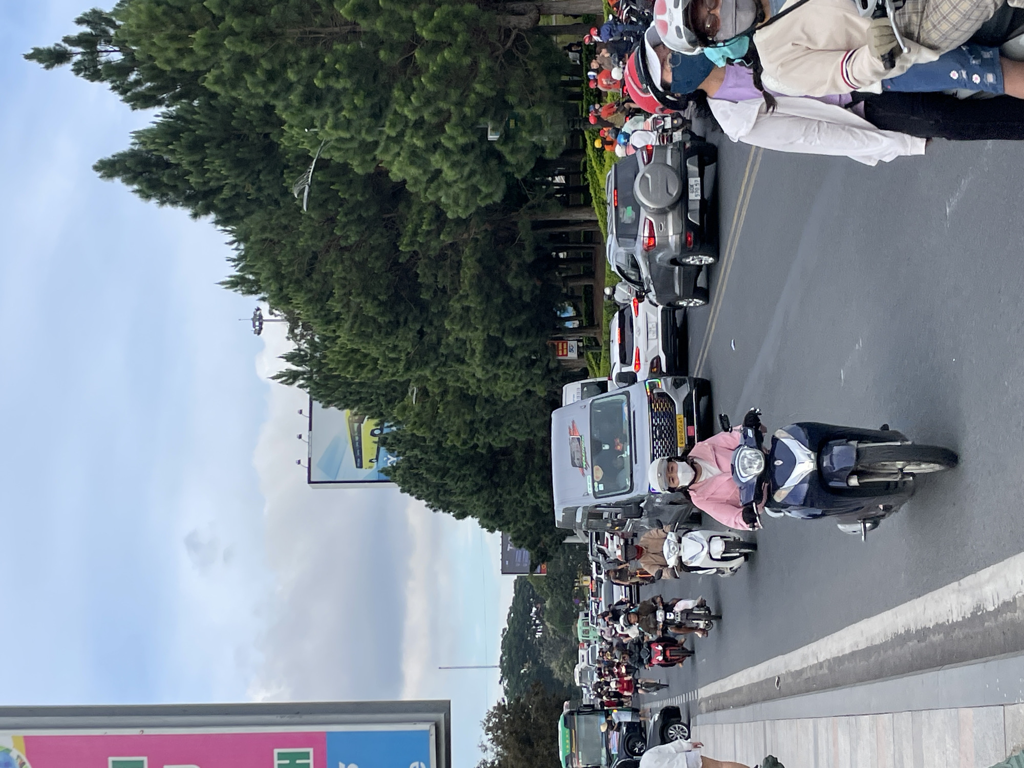 This image captures a busy street scene in Da Lat, Vietnam. A wide asphalt road is filled with a variety of vehicles, predominantly motorbikes, along with cars and vans. The traffic appears to be moderate to heavy, with vehicles moving in multiple lanes.
Several individuals are visible on motorbikes, most wearing helmets and face masks, a common sight in this region. One person in the foreground rides a dark blue motorbike, wearing a pink jacket and a white mask. To their right, another person on a motorbike wears a beige jacket and plaid shorts, also masked. In the mid-ground, other motorcyclists are seen, some with passengers. Cars and vans are interspersed within the traffic, including a silver SUV with a spare tire on the back and a grey minivan.
The environment suggests an urban or semi-urban setting. On the right side of the road, a dense line of tall pine trees forms a natural barrier, creating a green backdrop. Overhead, a cloudy sky is visible, with a hint of blue. In the background, a billboard with a yellow bus graphic is partially visible, and another large billboard with a pink and green advertisement is seen on the left edge of the frame. Streetlights and utility poles are also present, indicating a developed area. The overall impression is one of everyday life and movement in a Vietnamese city.