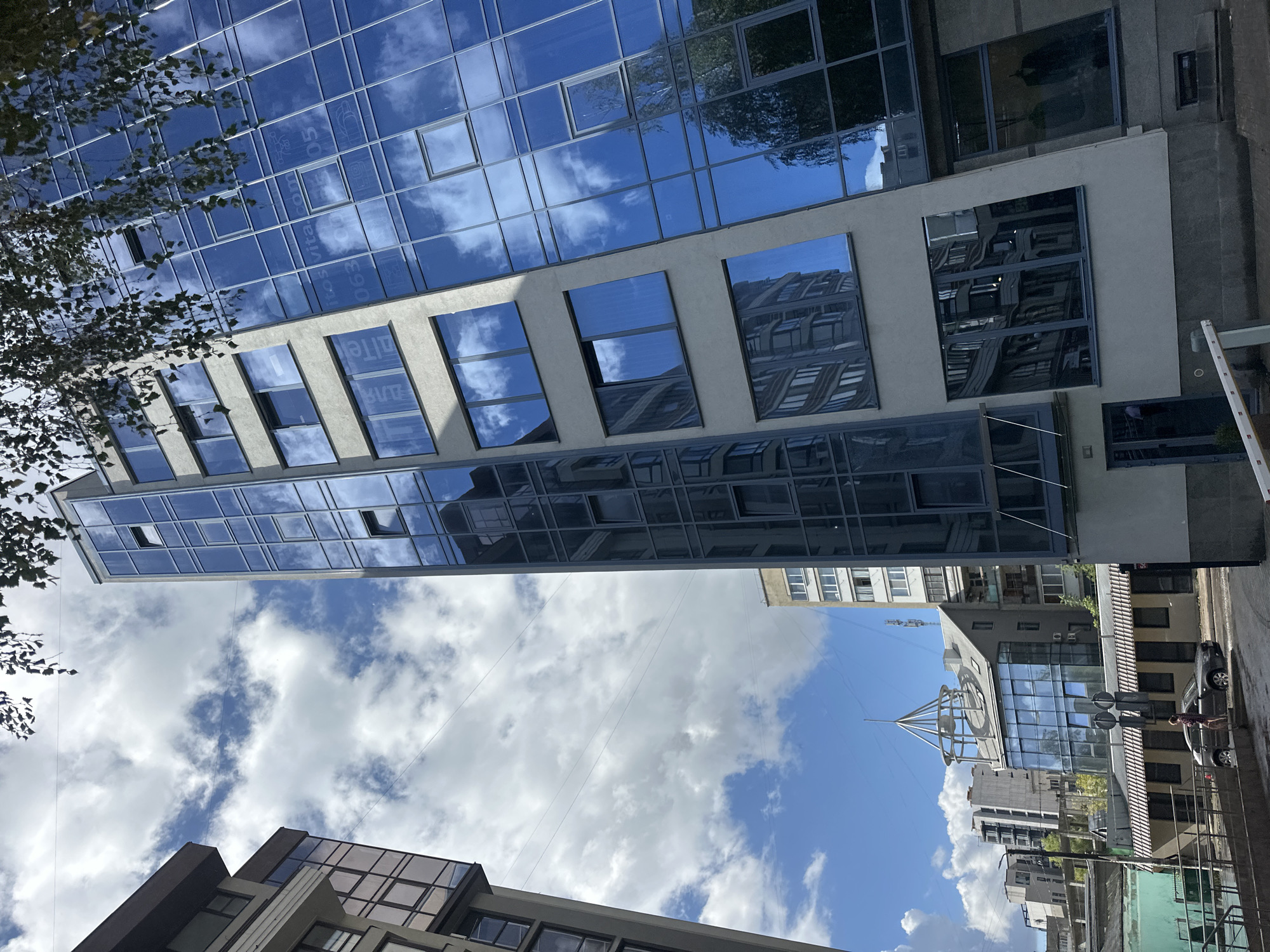 In Ternopil, Ukraine, a modern, multi-story building with a distinctive glass facade dominates the frame, angled from a low viewpoint. The building's blue-tinted glass reflects the cloudy sky and surrounding structures. Adjacent to it, another building with a lighter beige facade and dark-framed windows is visible on the left. In the background, a smaller building with a unique conical roof structure and glass walls stands out, surrounded by other urban constructions. A dark-colored car is parked near the curb in front of a low-rise building with a striped awning. A single person stands beside the car. The scene is set against a bright, partly cloudy sky, with tree branches from an unseen tree partially obscuring the top of the frame. Electrical wires are strung across the sky.