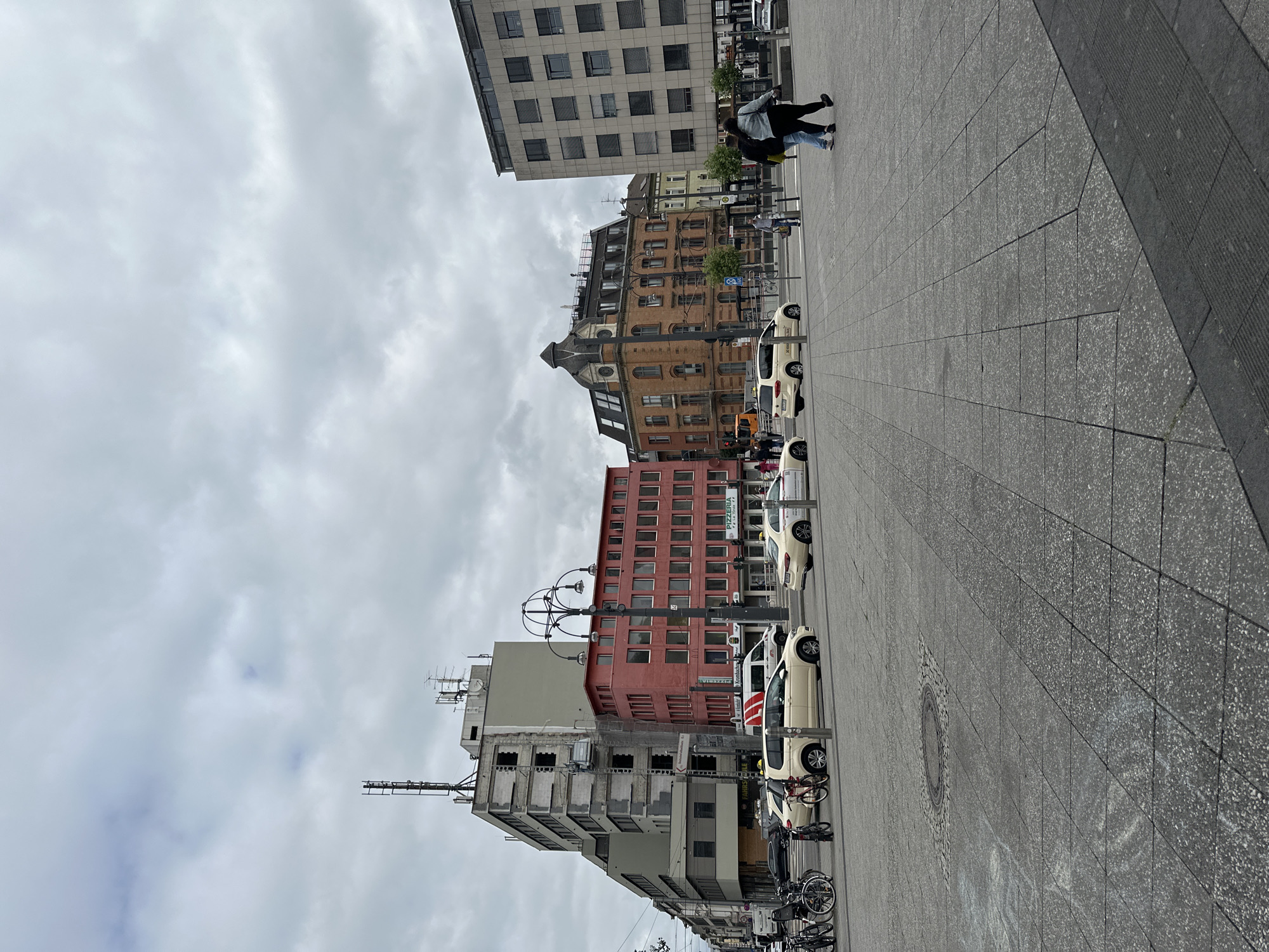 The image depicts a street scene in Krefeld, Germany, on an overcast day. In the foreground, a gray paved plaza with a manhole cover is visible. Several yellow taxis are parked or driving on the street. To the left, bicycles are parked near a building under construction or renovation, with scaffolding visible. Further back, a red building with numerous windows stands next to a taller, light gray building with a more modern design. To the right of the red building is an older, ornate brown brick building with a distinct mansard roof. The sky is filled with dense, gray clouds. Two individuals are walking away from the camera towards the right side of the frame. The scene suggests an urban environment with a mix of architectural styles and ongoing activity.