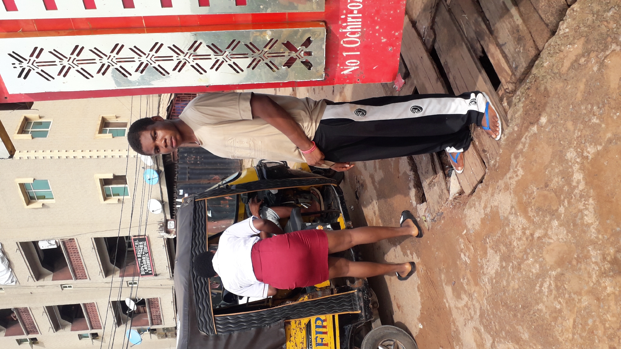 In Nkpor, Nigeria, a young male stands on wooden planks in front of a red sign that reads "No 1 Ochiri-Oz." He wears a beige t-shirt and black pants with white stripes. To his left, a woman in a white shirt and red skirt stands by the open door of a yellow and black auto-rickshaw. The woman is facing away from the camera, and her lower legs and feet, clad in black flip-flops, are visible. The auto-rickshaw has text on its side, including "DELUXE" and what appears to be "FIR." Behind the auto-rickshaw and the people, a multi-story building with balconies and windows is visible, along with various satellite dishes and electrical wires. The ground appears to be a mix of dirt and weathered wooden planks.