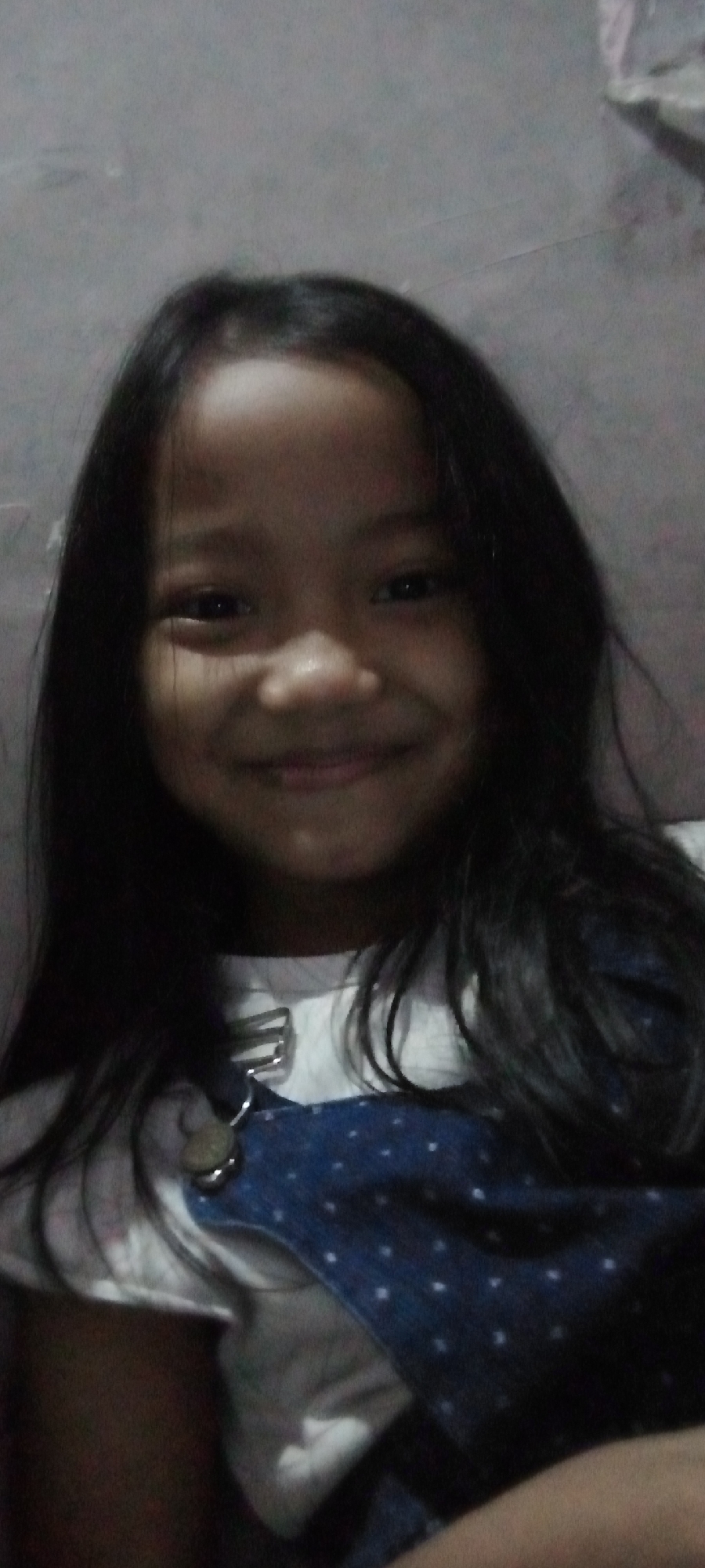 This is a medium shot, front-facing selfie of a young girl with long, dark hair. She has a broad smile and her eyes are dark. She is wearing a white t-shirt and a dark blue denim pinafore with small white polka dots. A silver clasp with a beige decorative element is visible on the pinafore strap. The background is a plain, gray wall, possibly in a home. The lighting appears to be artificial and somewhat dim. There are no other individuals or objects of note in the frame. The image quality is somewhat low resolution and grainy.