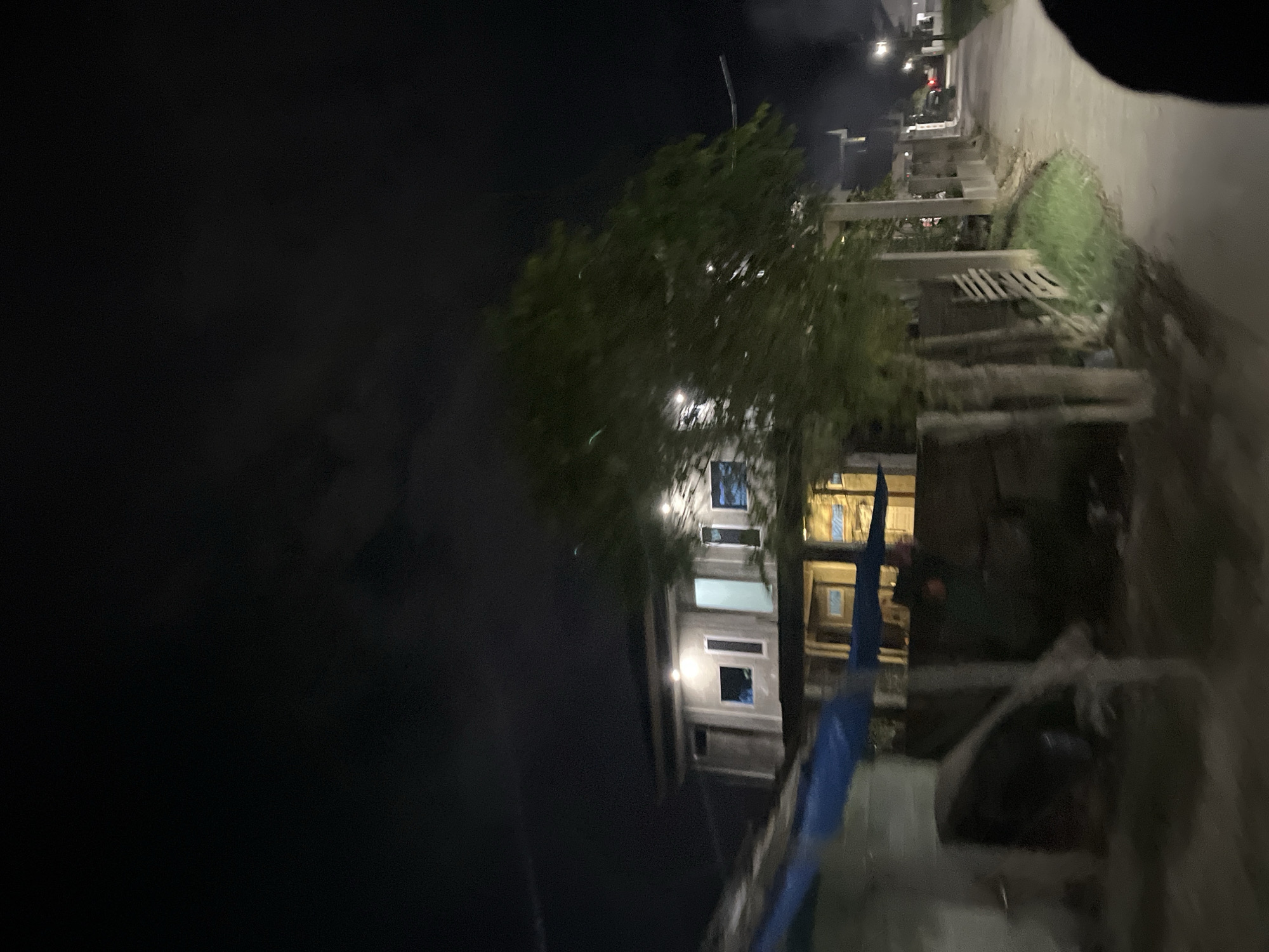 This media file captures a night scene in Menanggini, Indonesia. The image is oriented vertically, showing a street or pathway on the right side, bordered by a concrete wall and some vegetation. On the left, a dark, indistinct area dominates the frame, suggesting the night sky or a poorly lit area.
The central focus is a multi-story building with visible windows, some of which are illuminated, indicating interior lights are on. Trees with dense foliage are present, partially obscuring the building. The lighting is dim, consistent with nighttime, and there are faint lights visible in the distance, possibly from other buildings or streetlights. The overall atmosphere is dark and somewhat obscured, with a hint of the environment illuminated by artificial light. There are some visible construction materials or debris in the foreground, including what appears to be blue tarp. No people are clearly identifiable. The time of day is clearly night, and no specific weather conditions are evident. There is no visible text in the image.