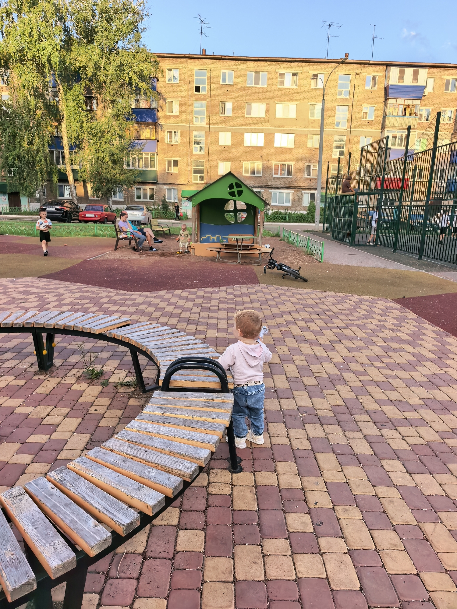 The image depicts a children's playground in Sterlitamak, Russia, adjacent to a brick apartment building. In the foreground, a young child wearing a pink hoodie and blue jeans stands facing away from the camera, holding a white object, possibly a bottle. The child is positioned next to a curved wooden bench. The playground features a colorful brick-patterned ground. In the middle ground, several people are visible. A young boy in a white t-shirt and black shorts walks across the playground, while two adults sit on a bench, and another child plays near a small green playhouse. A bicycle is propped up on the ground near the playhouse. The background shows a multi-story brick apartment building with numerous windows, and a chain-link fence surrounding a sports court. Lush green trees are on the left side of the image, and the sky is clear and blue.