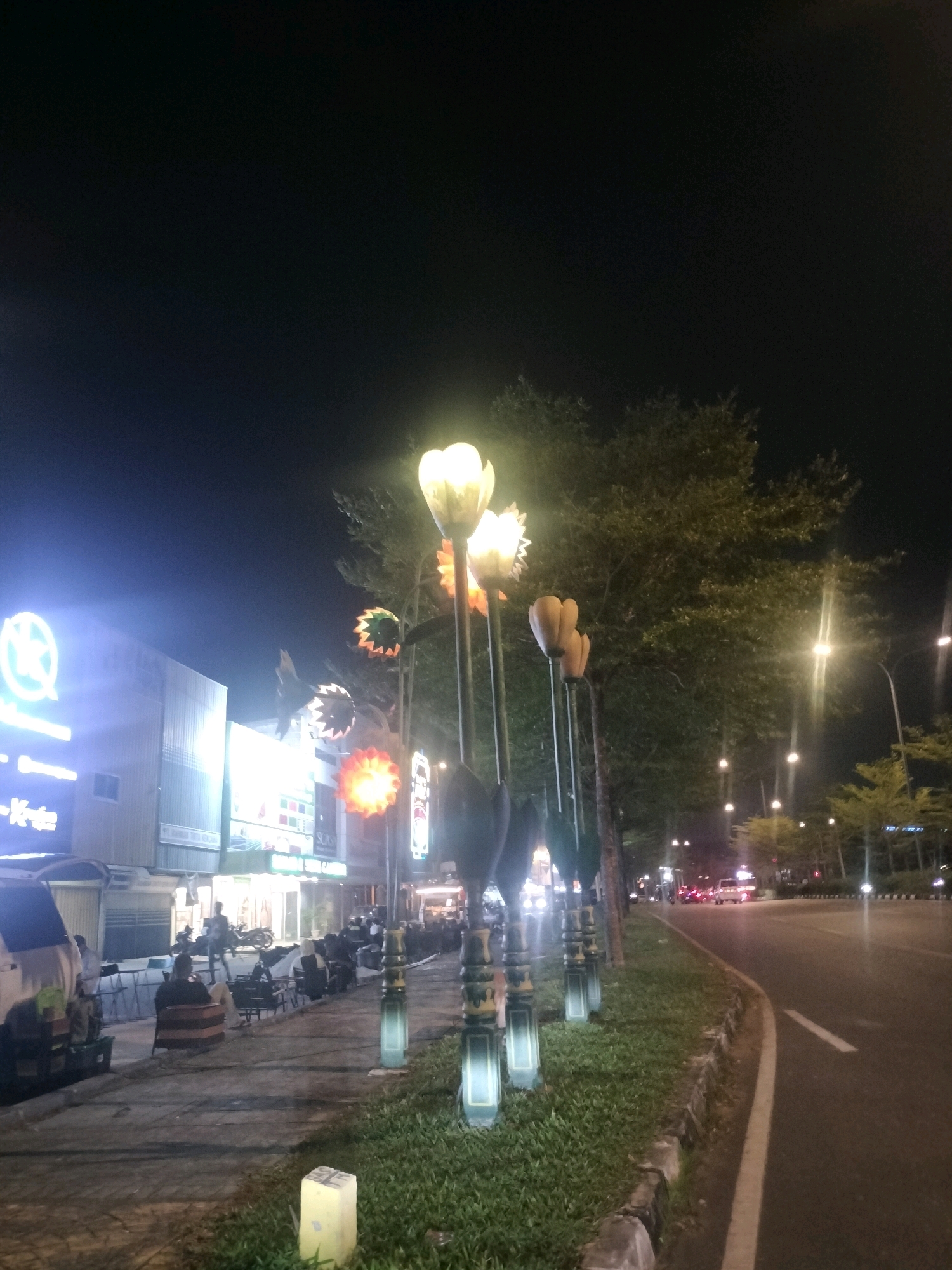 The image depicts a nighttime scene on a street in Batam, Indonesia. Several decorative lamps resembling large flowers are positioned along the sidewalk, illuminated with a warm, bright light. Behind the lamps, a large tree with dense foliage is visible, its branches extending upwards. Across the street, there are buildings with illuminated signage, suggesting a commercial area. The street itself is paved and appears to be a road with traffic, as evidenced by the headlights of passing vehicles in the distance. The sidewalk is lined with grass and stones, and there are people seated on chairs and benches, some appearing to be patrons of establishments. The overall atmosphere is one of a lively urban environment at night.