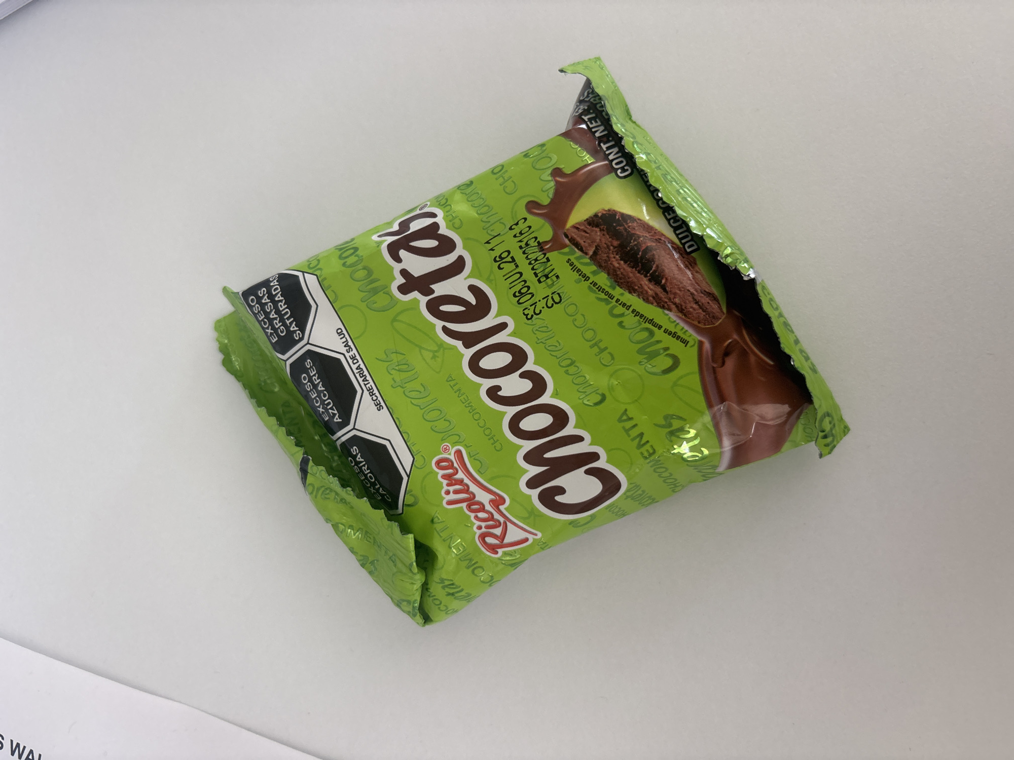 A single, unopened "Chocoretas" candy bar, produced by the Ricolino company, is diagonally positioned on a white surface. The candy wrapper is light green with "Chocoretas" printed in brown and numerous instances of "CHOCOMENTA" and "coretas" in green. A graphic shows a chocolate snack submerged in a chocolate liquid or sauce. A date of 06 JUL 26 is printed on the wrapper. Nutritional information, including warnings about excess calories, sugars, and saturated fats, is presented in a hexagonal design.
