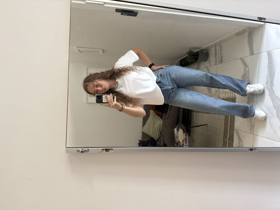 A young female individual is depicted taking a full-length mirror selfie within an indoor residential environment located in Tel Aviv-Yafo, Israel. The individual has long, curly light brown hair and is wearing a white short-sleeved t-shirt, light blue high-waisted jeans, and white sneakers. She holds a smartphone with a white and grey patterned case in her right hand, positioned to capture her reflection, while her left hand rests on her hip. Her attire is accessorized with a thin turquoise beaded necklace, several bracelets on her left wrist, and a black smartwatch also on her left wrist.
The mirrored reflection reveals a room with light-colored walls and a floor composed of white and grey marble-patterned tiles. To the left, a dark grey sofa is visible, adorned with a light green throw pillow and a laptop. A small, black, round coffee table with thin legs is positioned in front of the sofa, holding papers or magazines. To the right, a green skateboard leans against the wall next to a small potted plant housed in a grey pot. A white-framed object is mounted on the wall in the upper right portion of the reflection. The lighting is bright and ambient. The full-length mirror itself features a light metallic frame with visible hardware on its left side.