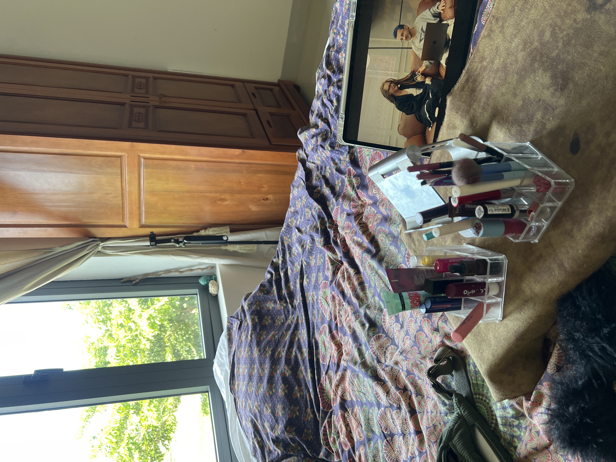 The image depicts an indoor scene, likely a bedroom, with a focus on a bed covered with a dark purple and pink patterned duvet. On the bed, towards the foreground, are two clear acrylic organizers filled with various makeup products, including lipsticks, brushes, and tubes of cosmetics. A laptop is also positioned on the bed, displaying a video call or streamed content featuring two individuals. One individual, appearing to be a woman with dreadlocks, is seated on a brown couch, while a man with blue hair is beside her, looking at the laptop.
To the left of the bed is a large window with a view of green foliage, suggesting an outdoor environment with trees. A light-colored curtain is drawn partially across the window. To the right of the window is a tall, wooden wardrobe with multiple doors and decorative paneling. The overall impression is a personal space, possibly a hotel room or a residential bedroom, in Đà Nẵng, Vietnam, given the context provided. The lighting suggests it is daytime.