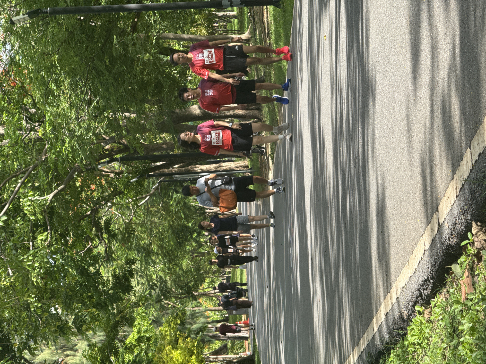 The image captures a shaded pathway in a park, likely in Bangkok, Thailand, given the lush greenery and tropical trees. The path is paved and marked with a yellow line along the edge, bordered by grass and foliage. Several people are walking or jogging along the path. In the foreground, a man in a grey t-shirt and black shorts is looking at his phone while holding an orange bag. Behind him, a group of three people in red t-shirts with race bibs are walking together, two men and a woman. Further down the path, other individuals and groups are visible, some appearing to be participating in a race or organized run. The scene is illuminated by sunlight filtering through the dense canopy of trees, creating dappled shadows on the path.