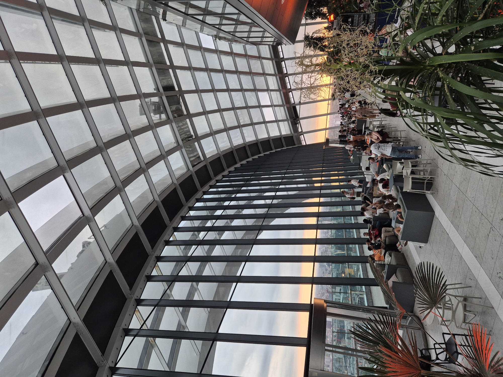 The image depicts the interior of a contemporary rooftop bar or restaurant in London, United Kingdom, with large glass windows offering a panoramic view of the city skyline at sunset. The architecture is modern and sleek, featuring a curved glass roof supported by dark metal beams, creating a spacious and open atmosphere.
Inside, there are multiple groups of people casually seated on modern lounge furniture, including sofas and chairs. Many are engaged in conversation, or looking out at the view. Some are standing, possibly socializing or waiting for drinks. The overall ambiance appears relaxed and social.
The foreground is occupied by lush green plants, adding a touch of nature to the urban setting. The lighting suggests it is either late afternoon or early evening, with the warm glow of the sunset casting a golden hue over the scene. The city below is visible through the expansive windows, showcasing a dense urban landscape with buildings and infrastructure. The presence of a bar area is also suggested in the background.
