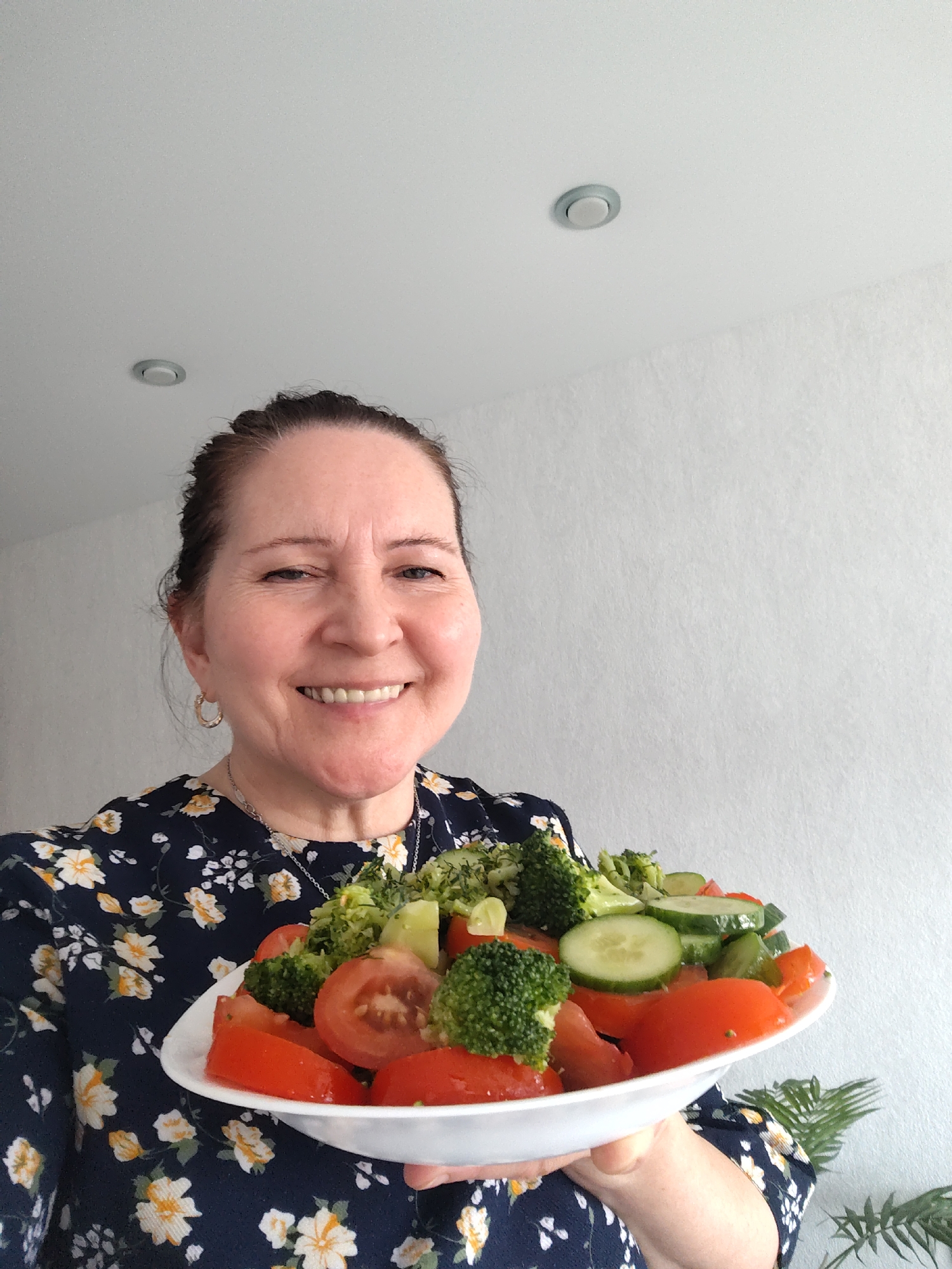 A smiling woman holds up a generous plate of fresh salad indoors. The woman, appearing middle-aged, has dark hair pulled back and wears a dark blue blouse adorned with a cheerful white and yellow floral pattern. She has a silver chain necklace and a gold-toned hoop earring on her left ear. Her face is turned towards the camera, radiating happiness with a wide, open-mouthed smile.
She presents a large white plate or shallow bowl filled with a vibrant, healthy salad. The salad prominently features bright red tomato wedges, numerous green broccoli florets, and sliced cucumbers. A faint glimpse of a green leafy plant is visible in the bottom right corner, behind her arm.
The background consists of plain light-colored walls, possibly white or light grey, displaying a subtle textured finish. A white ceiling is visible above, featuring a single recessed circular light fixture. The indoor setting suggests a domestic environment, potentially in Sarapul, Russia. The bright, even lighting indicates it is daytime. No text is visible in the image. The overall impression conveyed is one of health, contentment, and an appreciation for fresh food.