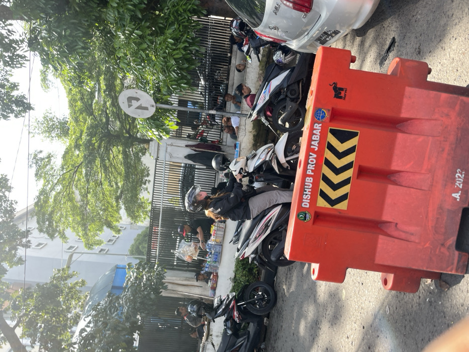 A woman wearing a helmet sits on a white and black motorcycle with a black and red motorcycle parked in front of her. Several people are standing and sitting on the sidewalk in the background, including a man with a helmet and a group of people wearing blue and red shirts. There is a large red barrier blocking the street, and a white car is parked behind the barrier. The road is paved and there is a large tree in the background. The scene is taken in the city of Bandung, Indonesia, at the coordinates -6.898 latitude and 107.597 longitude. The barrier is marked with the words "Dishub Prov Jabar" and the year 2022. There is a small elephant emblem on the barrier. The scene is a typical street scene in a busy urban environment in Indonesia.