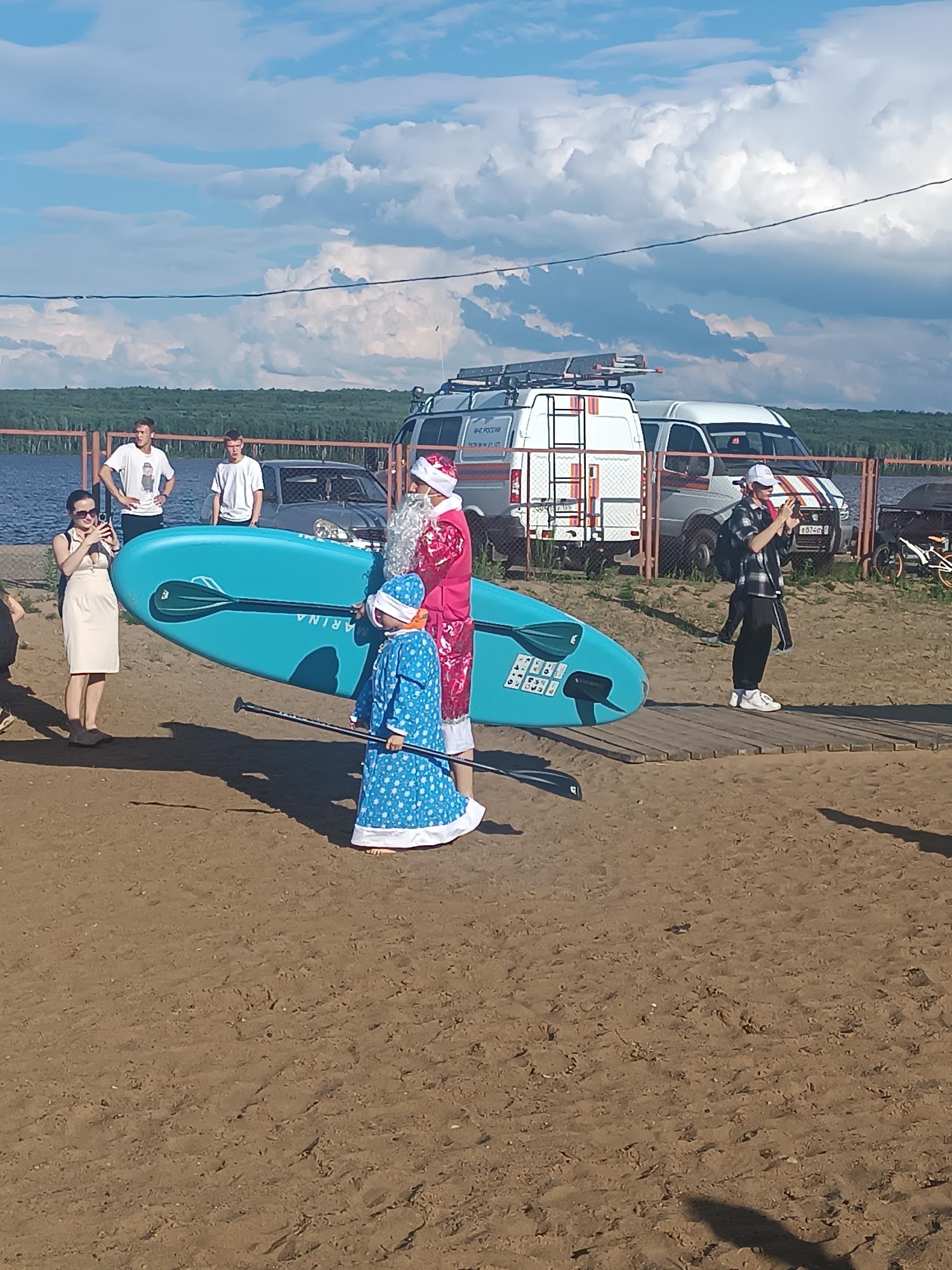 In Chaikovsky, Russia, a group of people are gathered on a sandy beach near a body of water. The sky is filled with large, dramatic clouds, suggesting partly cloudy or stormy weather. In the foreground, a woman in a light-colored dress stands holding a smartphone, seemingly taking a photo or video. Beside her, a blue paddleboard is held upright, with "ARINA" visible as text on its side.
A man dressed as Santa Claus, complete with a red suit and white beard, stands next to a child wearing a blue, ornate outfit. The child is also holding a paddle, and appears to be participating in the paddleboard activity or a festive event. Further back, two young men in t-shirts stand near a fence, and a few vehicles, including white vans and a car, are parked behind the fence, implying a parking area or event setup. The presence of Santa Claus and the child's festive attire suggest a holiday-themed event, possibly a summer celebration or a unique festive gathering in a warm climate. The overall mood seems relaxed and social.