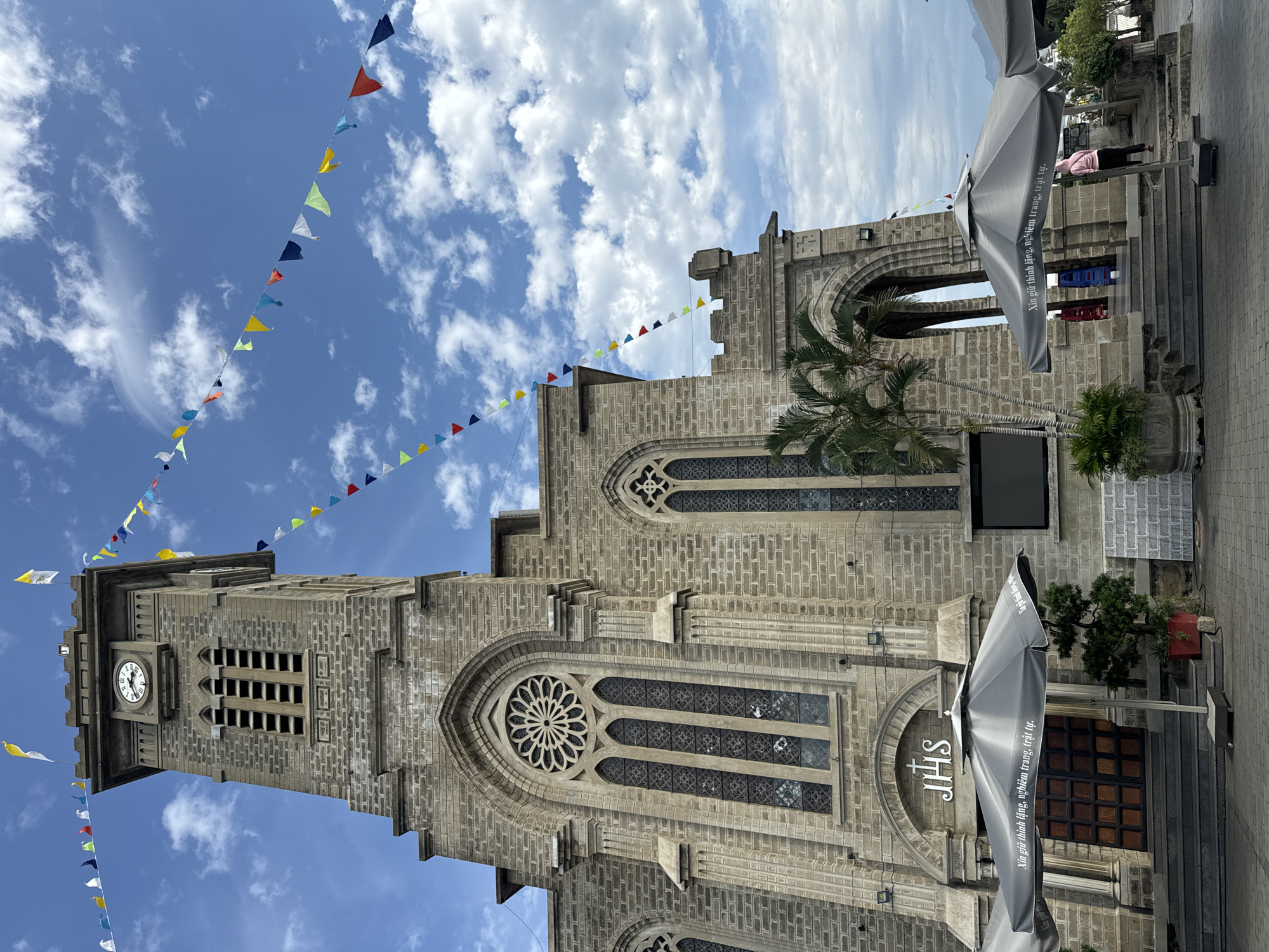 This is a vertical, eye-level shot of the Nha Trang Cathedral in Vietnam. The cathedral, constructed of grey stone with ornate architectural details, is the main subject. A tall bell tower with a clock face is visible on the left side of the image. Colorful triangular flags are strung across the bright blue sky filled with fluffy white clouds, adding a festive touch to the scene. Palm trees and other greenery surround the cathedral. In the foreground, a large, grey umbrella with text printed on it is angled towards the viewer. Part of a paved walkway is also visible. The text on the umbrella reads "Xin giữ thinh lặng, nghiêm trang, trật tự," which translates to "Please keep silent, solemn, and orderly." The overall impression is one of a sunny day at a significant religious landmark.