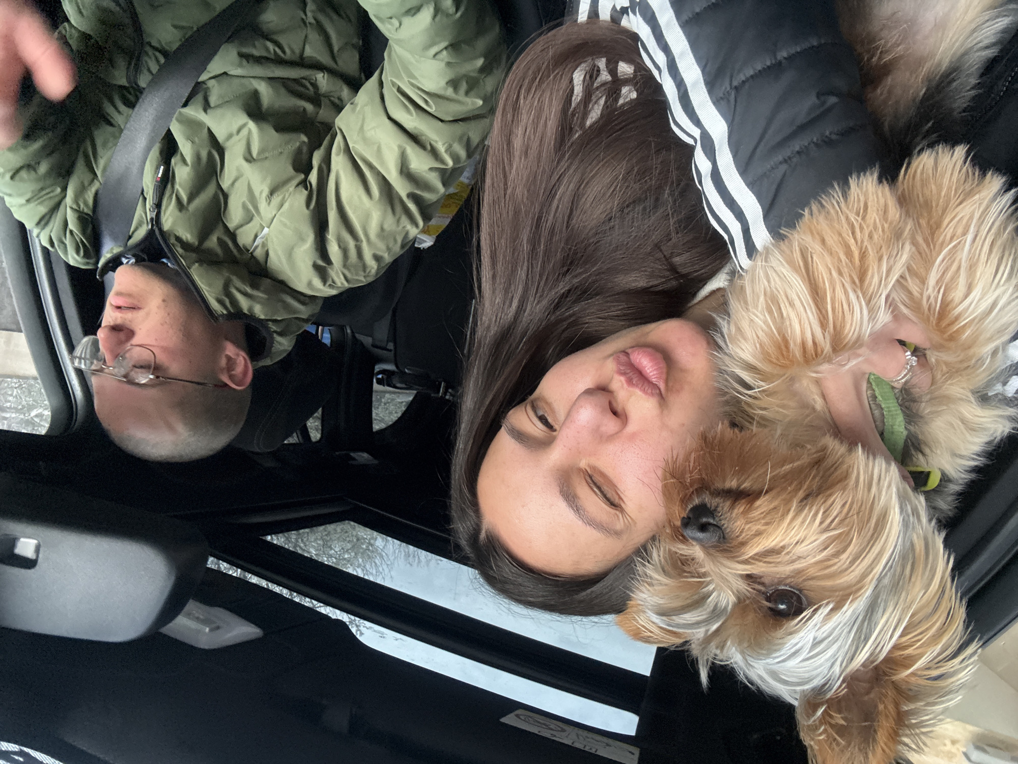 The image shows two people and a dog in a car, possibly a selfie taken by one of the occupants. The car has a sunroof.
One person is wearing glasses and a green puffer jacket with a black seat belt across their chest. The other person has long brown hair and is wearing a black jacket with white stripes. They are winking at the camera.
The dog is a small breed, possibly a Yorkshire Terrier, with long, light brown and white fur. It is being held by the person with brown hair.
The environment appears to be a car interior with a view of trees visible through the sunroof, suggesting an outdoor setting. There is no specific information to determine the location is in the city of Varna, Bulgaria.