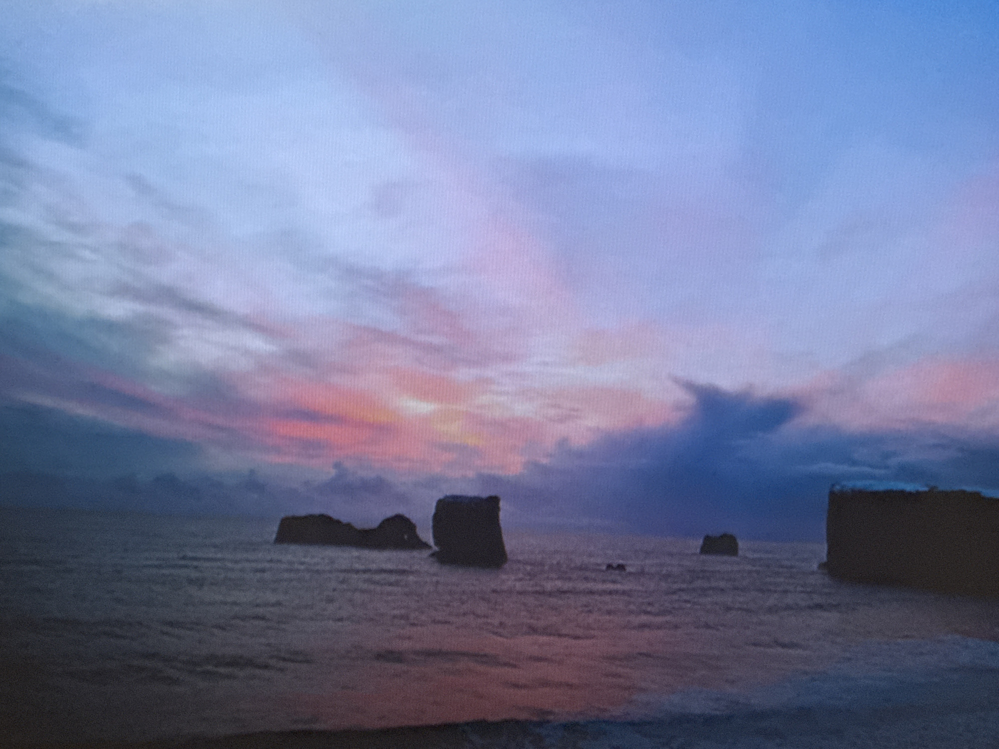 A wide shot captures a dramatic dusk sky over a serene body of water. The sky transitions from a soft, pale blue at the top to hues of pink and purple as it descends, painted with wispy clouds that radiate outwards. The water below reflects the sky's colors, with gentle ripples disturbing the surface. In the middle ground, several dark, silhouetted rock formations emerge from the sea, their shapes stark against the soft light. One prominent formation stands tall and rectangular on the right side of the frame, while others are more scattered and irregular. The shoreline in the foreground is dark and indistinct. There are no people or any man-made structures visible in this image.