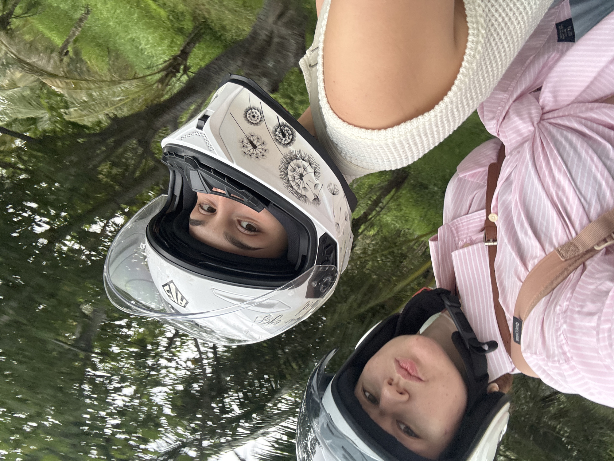 Two individuals wearing motorcycle helmets are positioned closely together in an outdoor environment with lush green foliage and trees in the background, suggesting a tropical location such as Rawai, Thailand. The individual in the foreground wears a black and gray helmet and a pink and white striped shirt, with a brown leather strap across their chest. Their gaze is directed towards the camera with a slightly pursed expression. The individual behind them wears a white helmet adorned with a dandelion-like graphic and is looking upwards, with their right eye visible through the helmet's visor. Their upper body is clad in a cream-colored knitted top. The scene appears to be captured from a moving vehicle, possibly a motorcycle, given the tilt of the image and the framing of the individuals. The overall atmosphere is casual and dynamic, suggesting a shared activity or journey in a natural setting.