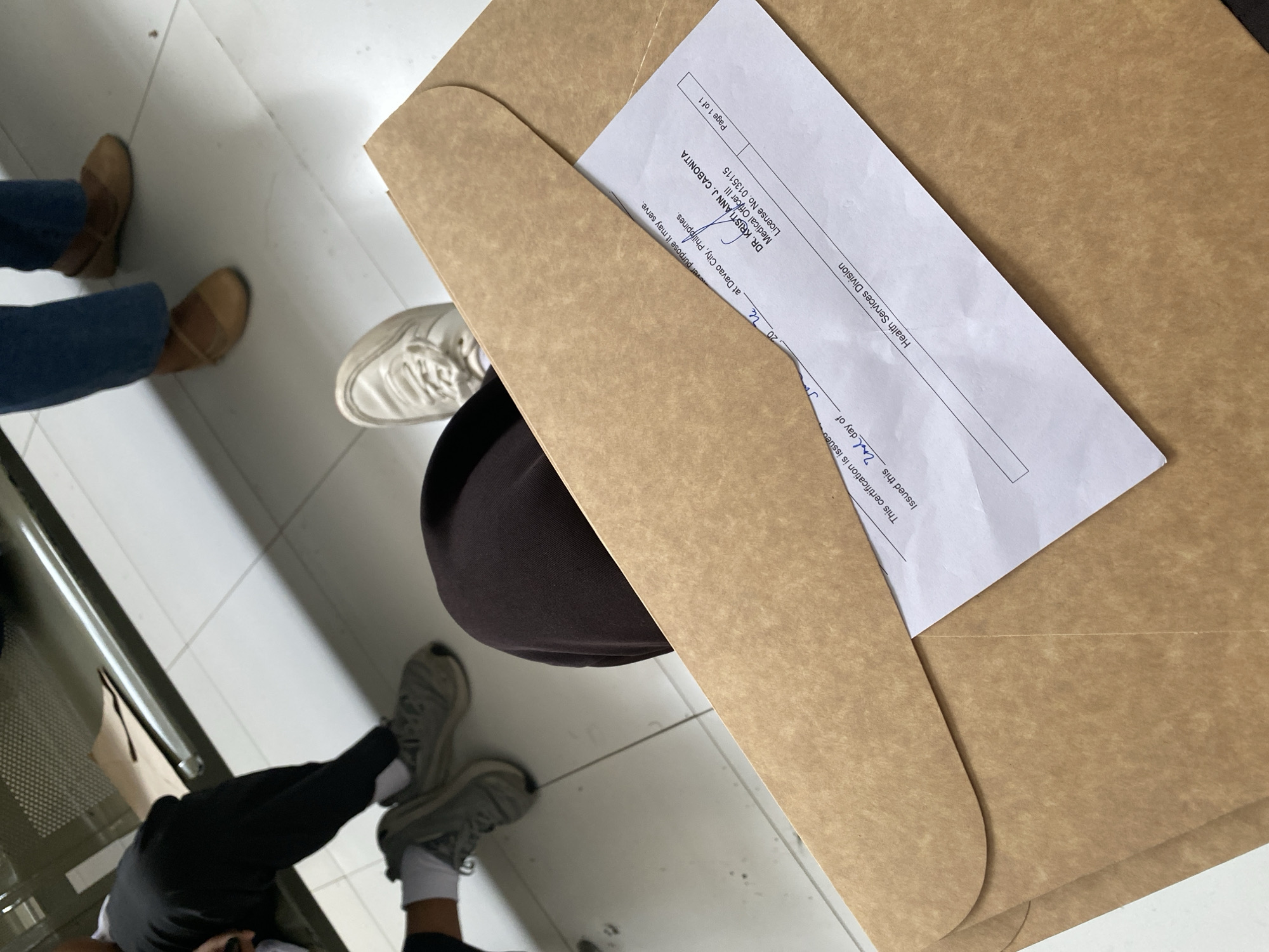 A close-up, overhead shot shows a person's legs and feet, one in black pants and gray sneakers, the other in dark brown pants and white sneakers, sitting with a large brown envelope on their lap. The envelope contains a document with visible text. The document shows a signature and the name "DR. KRISTIANN J. CABONITA," along with "Medical Officer III" and "License No. 0135115." It also mentions "Davao City, Philippines" and "Health Services Division." The background reveals a tiled floor and a metal bench, suggesting an indoor waiting area, likely in Davao City, Philippines.