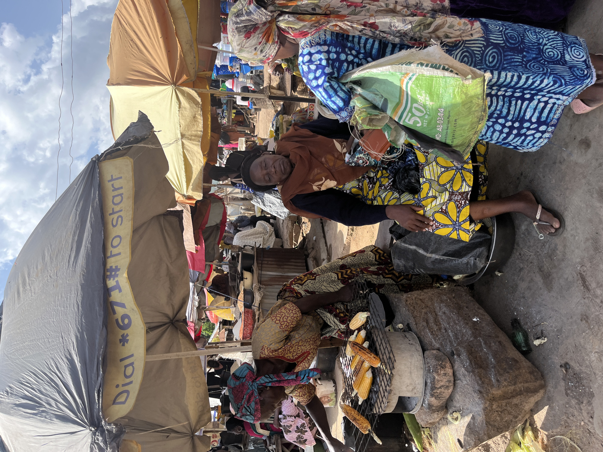 The image depicts a vibrant outdoor market scene in Gwong, Nigeria. In the foreground, two women are engaged in selling and preparing food. One woman, wearing a colorful yellow and black patterned dress, is seated and appears to be resting. The other woman, in the lower half of the image, is tending to corn being grilled over an open fire. She wears a patterned headscarf and a dress with a geometric design. The grilling setup includes a metal rack over what appears to be a makeshift stove or brazier, with several ears of corn cooking.
Behind the women, the market unfolds with various stalls and vendors. Numerous umbrellas and awnings provide shade. One large umbrella, predominantly gray and khaki-colored, has a yellow band with purple text. Partially visible text on this band reads "Dial *671#. To S". Another green bag, with visible text "50kg NET WEIGHT" and a phone number, is placed near the seated woman.
The scene is bustling with activity, suggested by the presence of many people and a variety of goods on display, though not all are clearly discernible. The lighting indicates it is daytime, likely with sunny weather given the strong shadows and bright sky visible in the upper left portion of the image. The overall impression is one of lively commerce and everyday life in a Nigerian market.