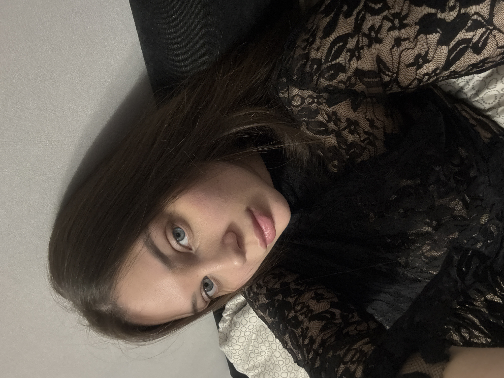 A woman with light skin, blue eyes, and brown hair is lying down. She is wearing a black lace top. The background includes a gray wall and a black object, possibly a piece of furniture. The context suggests that the photo was taken in an indoor environment. There is no evidence to confirm the location is Chaikovsky, Russia.