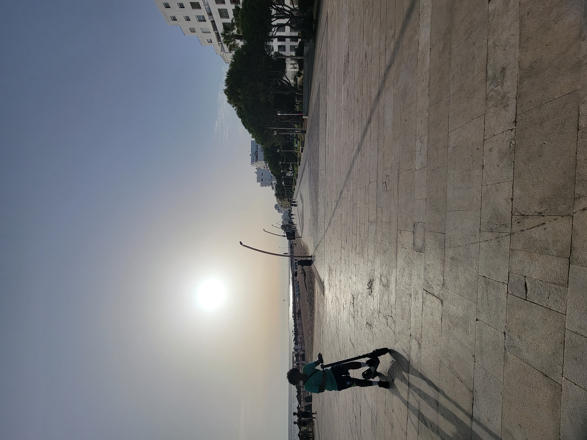 The image depicts a sunny day at the Quarteira waterfront in Portugal. In the foreground, a person is standing on an electric scooter on a paved promenade. The person is silhouetted against the bright sun, wearing a teal t-shirt and dark shorts. The promenade is made of large, light-colored rectangular stone slabs, and long shadows are cast across it.
To the left of the promenade, a sandy beach is visible with beach umbrellas and some people walking. The ocean stretches to the horizon, with calm water reflecting the sunlight.
To the right of the promenade, a line of white buildings with multiple floors and windows runs along the coastline, interspersed with trees and lampposts. The architecture suggests a resort or residential area. The sky is a clear, light blue with the sun prominently positioned in the upper left quadrant of the frame. The scene conveys a peaceful, recreational atmosphere typical of a seaside town.