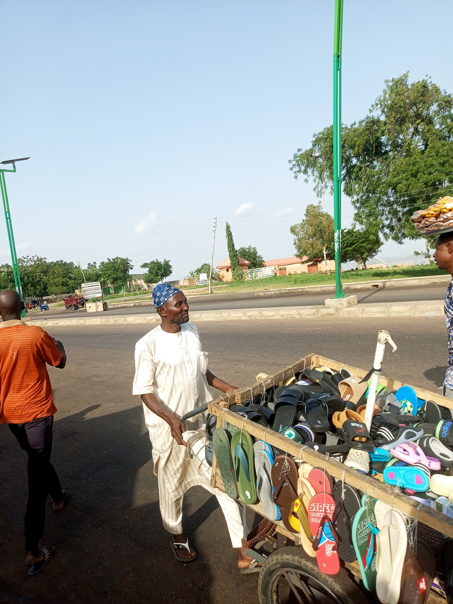The image depicts a street scene in Gombe, Nigeria, with two men and a cart filled with footwear. In the foreground, a man wearing a white, striped tunic and a blue and white patterned head covering is pushing a wooden cart laden with a variety of flip-flops and sandals. The footwear, in numerous colors and designs, is displayed on multiple levels, some hanging from the sides of the cart. To the left, another man in an orange, striped t-shirt and dark trousers walks away from the camera, his back to the viewer. He is wearing flip-flops. The cart has a single wheel visible at the bottom right. In the background, a road runs horizontally, with trees and buildings lining the opposite side. A lamppost with solar panels stands prominently on the left side of the image. The sky is clear and blue.