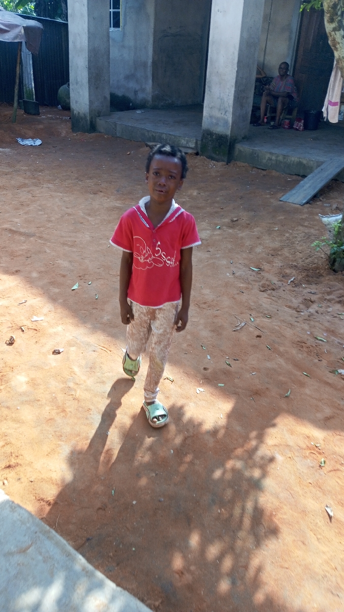 A young girl stands in the center of the image, wearing a red t-shirt, patterned pants, and sandals. She is on a dirt path in front of a concrete building with a seated man visible on its porch. Sunlight casts shadows of trees and the girl onto the ground. The scene suggests a rural or residential setting.