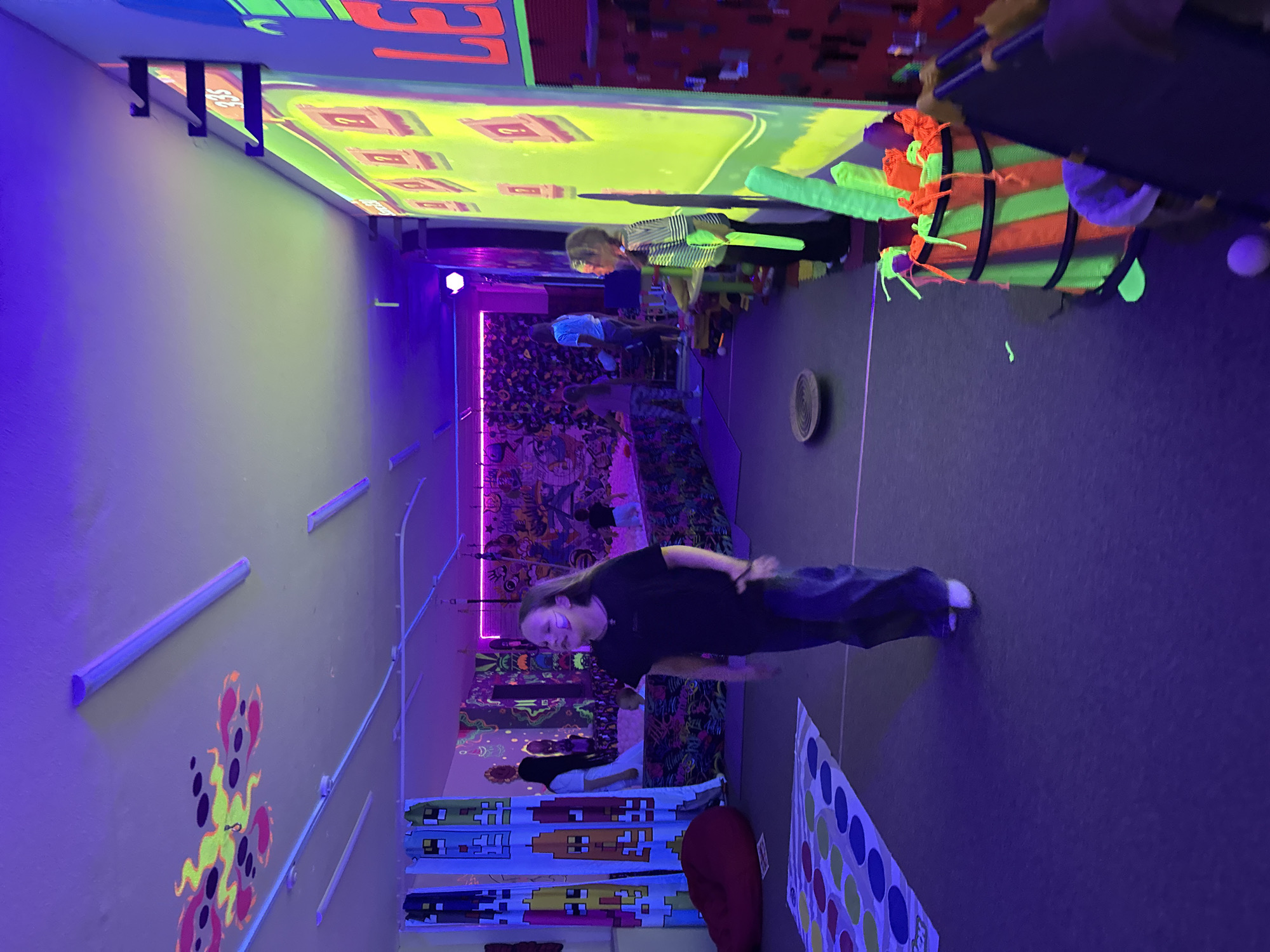 A group of young people are engaged in activities within a room illuminated by blacklights and neon lights. The scene is vibrant and colorful, with neon green, pink, and blue lights creating a glowing ambiance.
In the foreground, a person with long hair, wearing a black t-shirt and jeans, is captured mid-motion, possibly dancing or playing a game. Their face shows a smile, suggesting enjoyment. To the right of this individual, a Twister mat is laid out on the floor, hinting at a game in progress.
Further back, other individuals are visible. One person in a striped shirt appears to be interacting with a projection on the ceiling, which displays brightly colored squares and shapes. Another person in a white shirt is seated.
The walls are decorated with various colorful designs, some of which appear to be glowing under the blacklights. There are also what look like projection screens displaying abstract patterns or game elements. The overall setting seems to be an entertainment or activity center designed for a fun, lively experience. The time of day is not discernible, but the artificial lighting suggests an indoor environment.