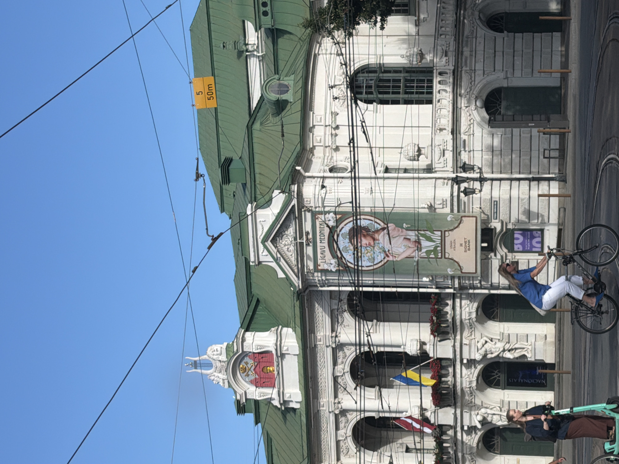 The image depicts the exterior of the Latvian National Opera and Ballet building in Riga, Latvia. The building features white stucco facade with ornate architectural details, a green roof, and large arched windows. In front of the building, there are tram tracks and people on a street. One woman is riding a bicycle, and another is standing on an electric scooter. Two flags, the Latvian flag and the Ukrainian flag, are visible, as well as a large banner with a painting of a woman. A yellow sign with the number "5" and "50m" is attached to the roof. The sky is clear and blue.