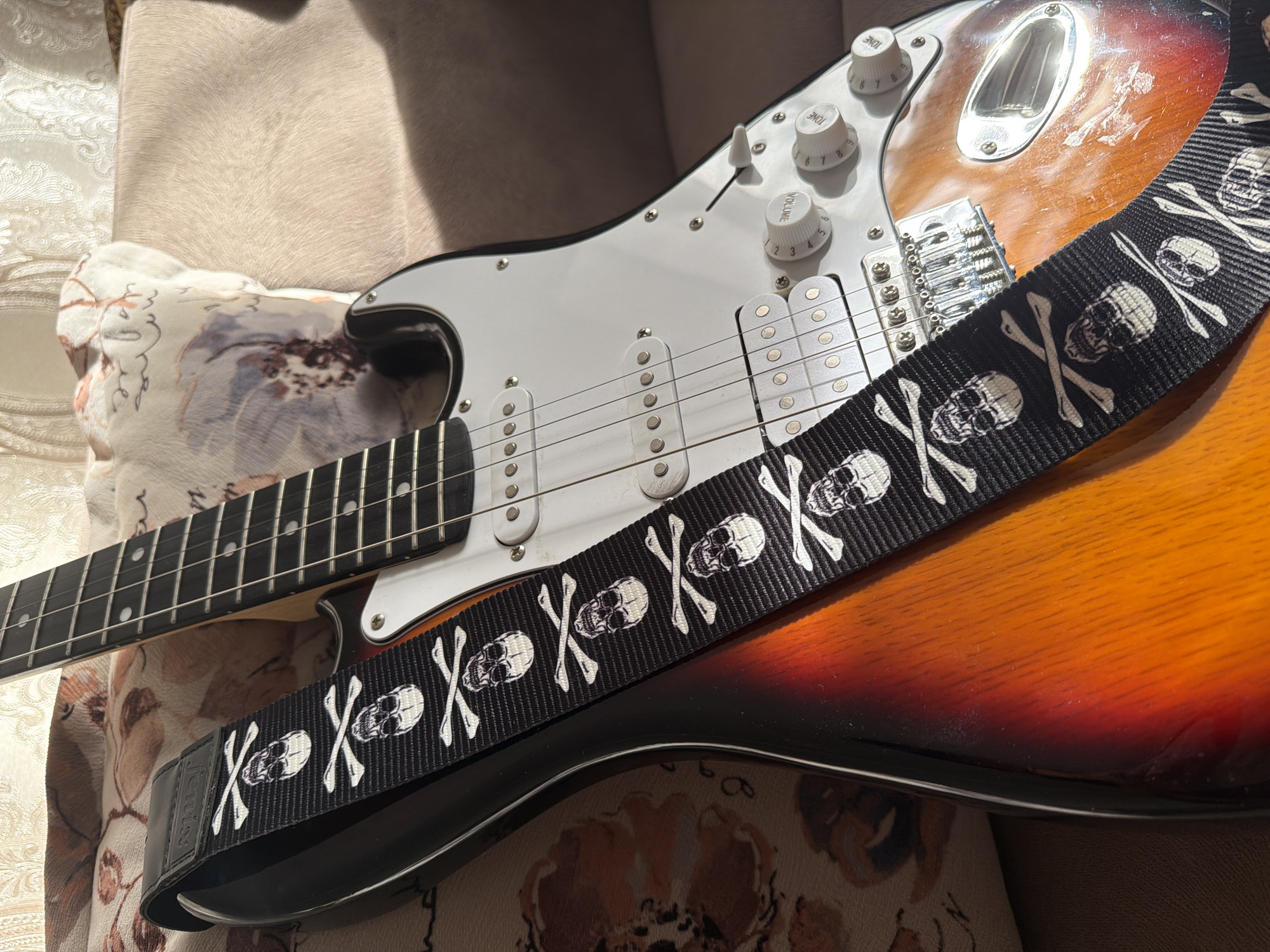 The image features a close-up of an electric guitar, resting on a patterned fabric surface, possibly a sofa or cushion. The guitar has a sunburst finish, transitioning from brown to orange. A black guitar strap, adorned with a repeating pattern of white skulls and crossbones, is draped across the body of the guitar. The guitar's white pickguard is visible, along with its chrome hardware, including tuning pegs, pickups, and a bridge. The strings of the guitar are also clearly visible. The lighting suggests it is daytime, with shadows indicating natural light casting onto the scene. The overall impression is one of a musician's instrument, possibly in a home setting. There is no visible text.