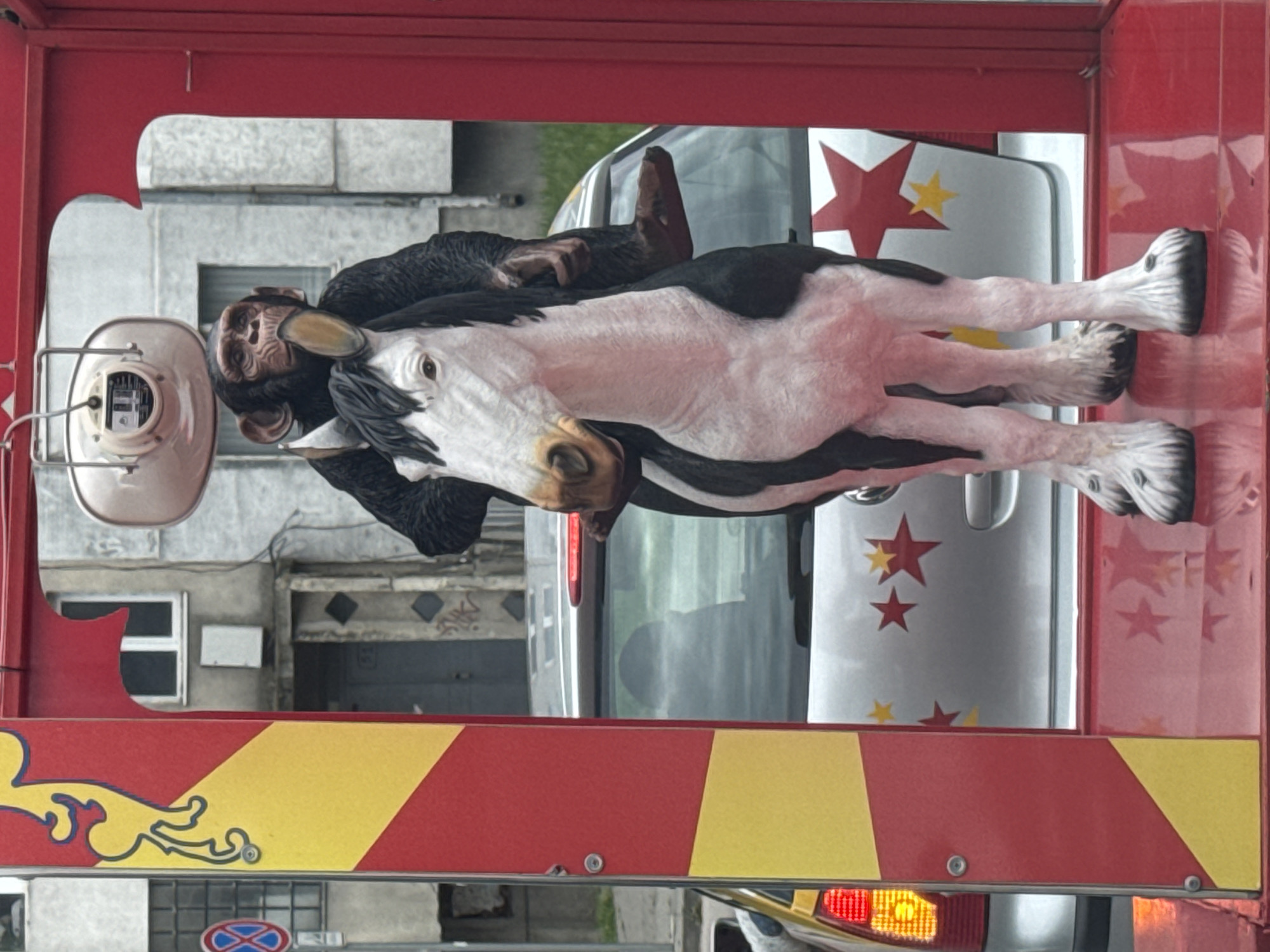 A surreal scene is captured: a chimpanzee figure rides atop a model horse, both perched on a trailer adorned with red and yellow stripes. The trailer is parked on a street in Varna, Bulgaria, as evidenced by a road sign visible in the background. Behind the trailer, a silver vehicle is partially visible, decorated with star motifs. Buildings are situated behind the vehicles, presenting an urban backdrop.