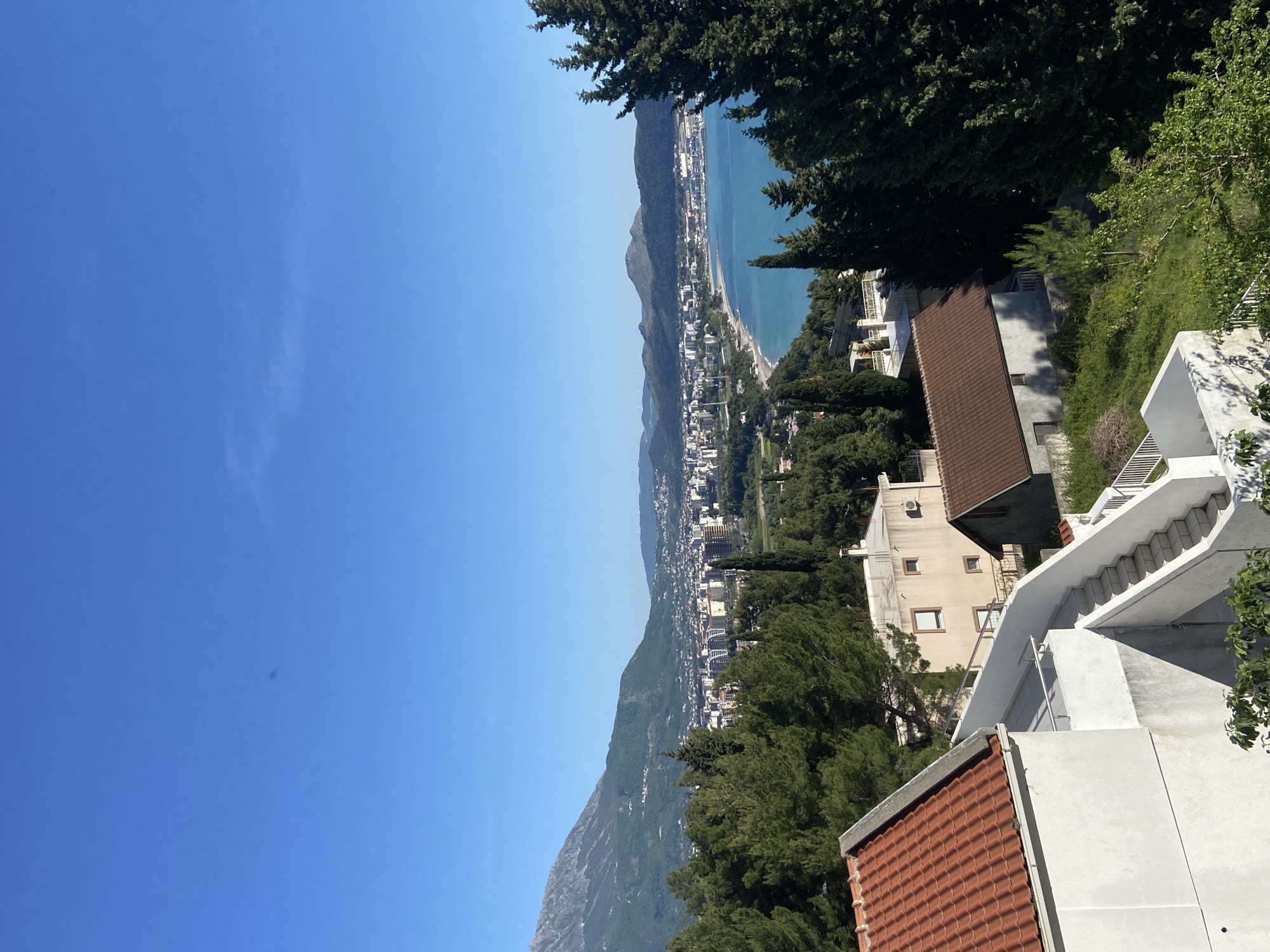 This image captures a panoramic, high-angle view of the coastal town of Šušanj, Montenegro, under a clear blue sky. The perspective looks down upon the Adriatic Sea and the sprawling settlement nestled between the water and the base of majestic mountains.
The top portion of the image is dominated by an expanse of vibrant, cloudless blue sky, suggesting bright, sunny weather, likely midday or early afternoon given the distinct shadows. The sea, a deep turquoise, stretches along the coastline, meeting a long beach that parallels the town. The town itself features numerous white and light-colored buildings with red-tiled roofs, densely populating the narrow strip of land between the sea and the foothills. A prominent mountain range rises sharply from behind the town, its slopes covered in greenery and some visible rock formations.
In the foreground, on the right side of the frame, several residential buildings with typical Mediterranean architecture are visible. These multi-story structures have white walls and distinctive terracotta-colored tiled roofs. Intricate white external staircases are a prominent feature, connecting different levels or parts of the buildings. Lush green trees, including tall conifers and deciduous varieties, are abundant, framing the scene and scattered throughout the landscape. One building in the mid-ground shows an air conditioning unit on its exterior and a ladder leaning against a wall.
No people are visible, and there is no discernible text in the image. The scene conveys a peaceful, picturesque coastal landscape under optimal weather conditions.