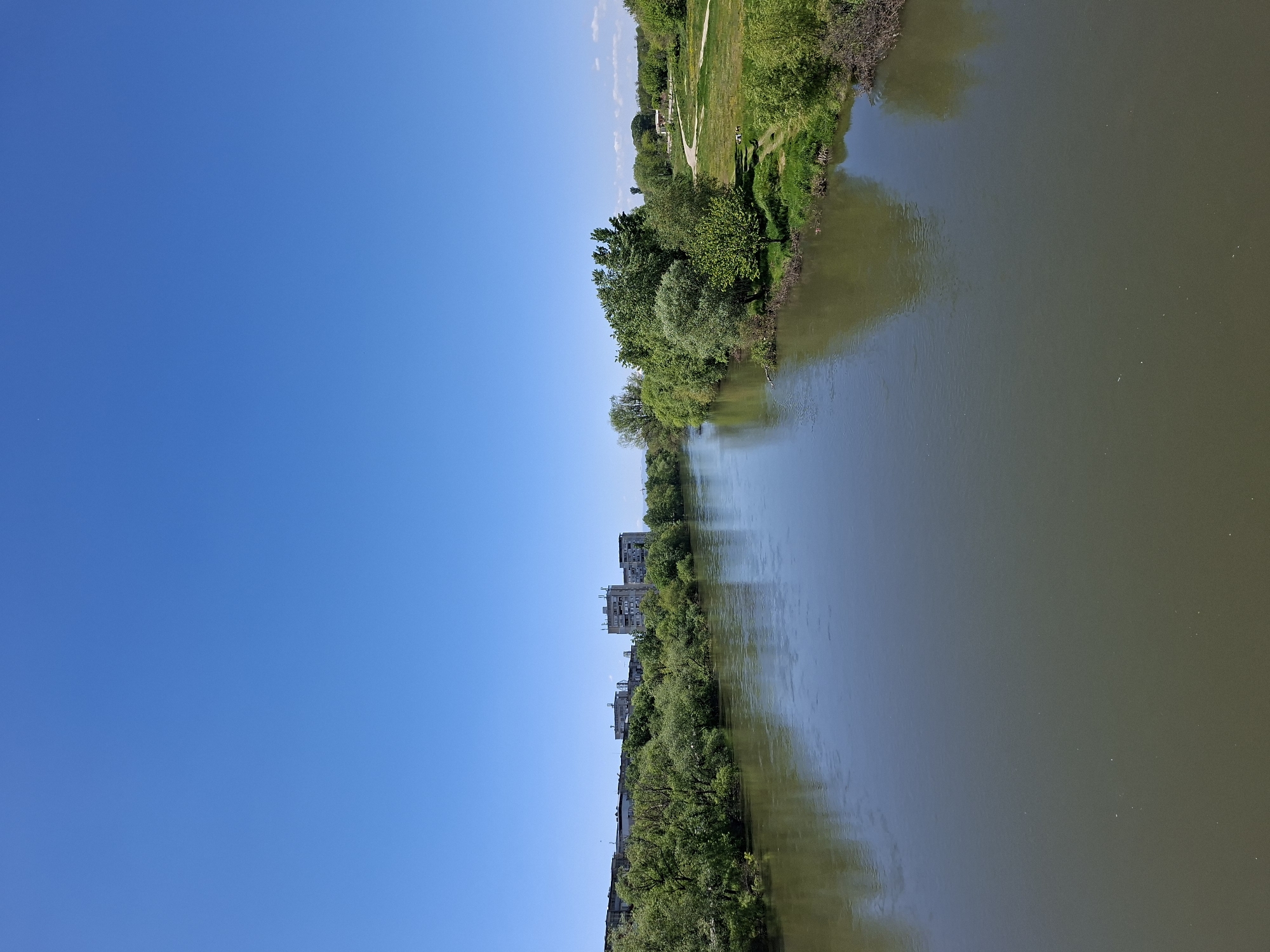 The image shows a river running through Plovdiv, Bulgaria. On the right side of the river, there is lush green vegetation, including trees and grass. The river itself is a murky color. On the left side, there are buildings visible among the trees. The sky is clear and blue. The image is taken from a high vantage point.
