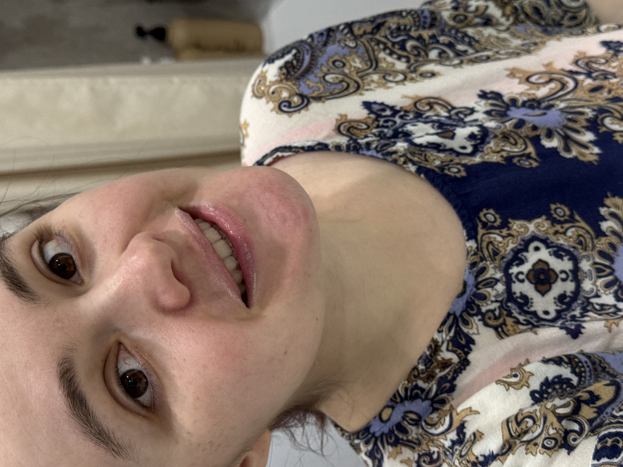 A person, presumably female, is shown in close-up. She has light skin, brown eyes, and dark eyebrows. She is smiling slightly, revealing her teeth. She is wearing a blouse with a blue, white, and gold paisley-like print. The background is blurred, with a light-colored surface visible behind her head. The location of Izhevsk, Russia is not discernible from the provided image.